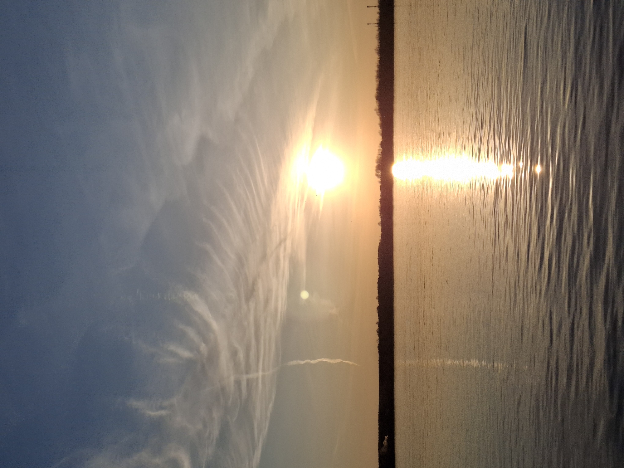 The image captures a serene sunset or sunrise over a vast body of water, likely a lake or wide river, with a distant treeline or low-lying land forming the horizon. The sky, occupying the upper left portion of the image, displays a gradient of blues and grays, transitioning to warm golden and orange hues closer to the sun. Wispy, elongated clouds stretch across the sky, catching the light and exhibiting intricate patterns, particularly on the left side of the frame.
The sun is a brilliant, blinding orb positioned centrally in the upper half of the image, casting a powerful golden glow. Its light illuminates the sky around it and creates a striking reflection on the water's surface. Below the sun, a vibrant, shimmering path of sunlight extends across the water towards the foreground, composed of numerous bright specular highlights on the rippling surface. The water itself, which fills the right side of the image, appears calm but with subtle waves or ripples, especially evident in the way the sunlight reflects off it.
A dark, thin silhouette of a distant shoreline or landmass runs horizontally across the mid-section of the image, separating the sky from the water. Some faint, slender vertical structures, possibly trees or utility poles, are barely discernible along this horizon line. The overall atmosphere is peaceful and natural, emphasizing the beauty of golden hour light over water. There is no visible text in the image.