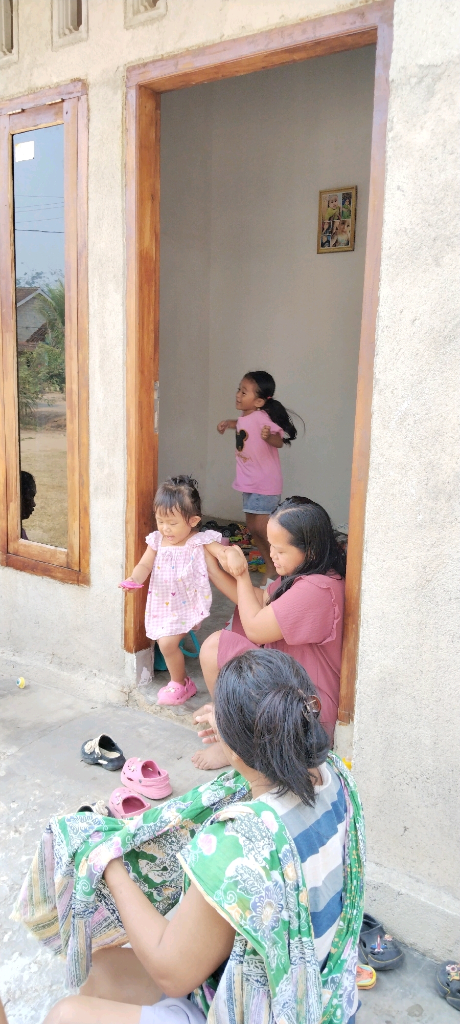 In Tanjung Ratu Ilir, Indonesia, an outdoor scene unfolds in front of a simple building with a wooden door frame. Inside the doorway, a young girl in a pink dress with a cartoon character on it stands on a raised platform, her arms outstretched as if dancing. She has dark hair pulled back into a ponytail. Nearby, a toddler in a pink and white checkered dress and pink Croc shoes stands, reaching out to a woman kneeling on the ground. The woman, wearing a dusty rose-colored dress, is holding the toddler's hands. She has dark hair pulled back and appears to be interacting with the child. In the foreground, another woman is seated on the ground, facing away from the camera. She is wearing a green and white patterned cloth draped over her and a striped shirt. Her dark hair is pulled back. Scattered on the ground are several pairs of children's shoes, including black and pink Crocs. A framed picture with multiple smaller photos is visible on the wall inside the doorway. The building's exterior wall is a rough, light-colored concrete. A window with wooden frames is to the left of the doorway, reflecting the outdoor environment, including a house and trees. The ground is concrete.