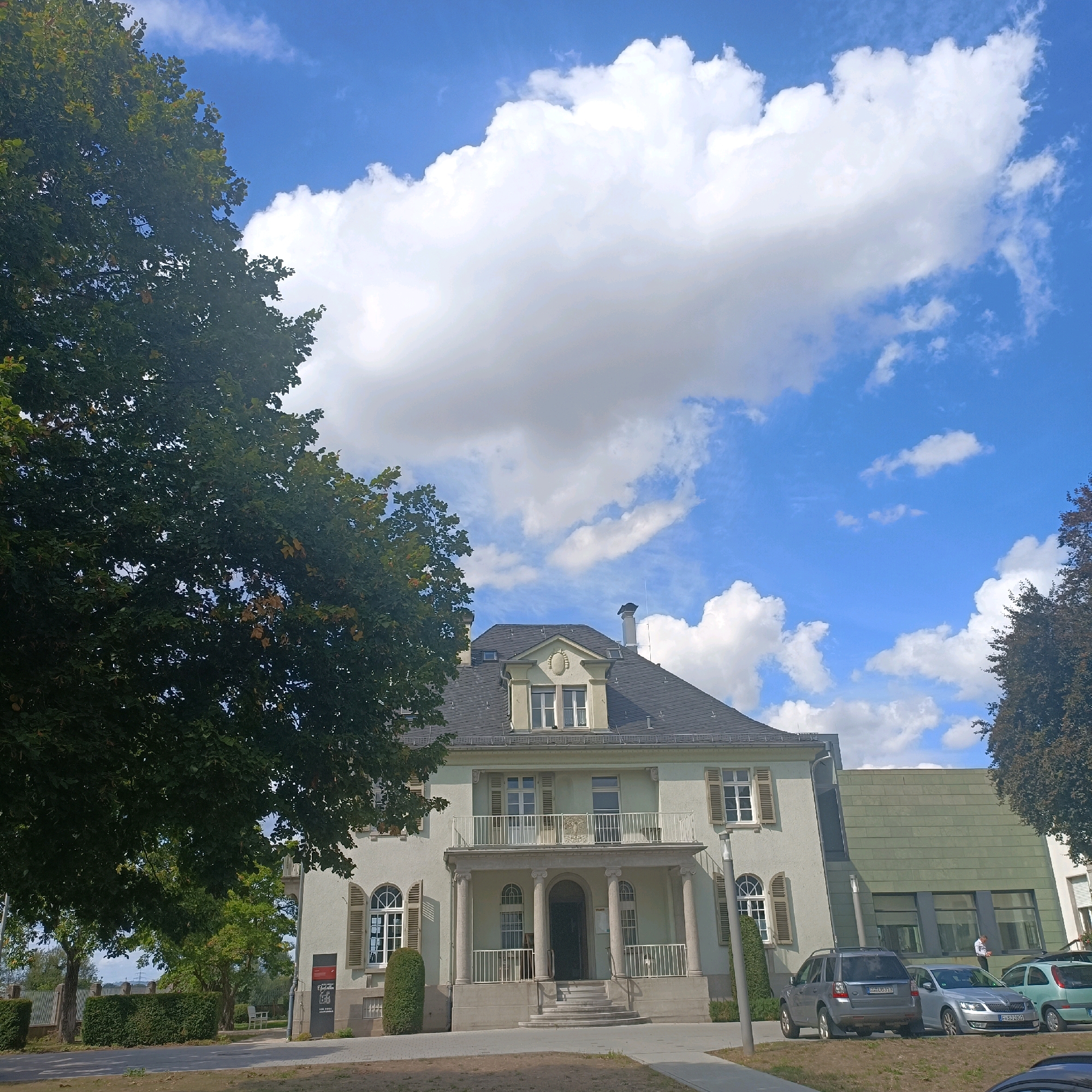 This image depicts a large, light-colored building with a grey roof, situated in a suburban setting. The building features a prominent central entrance with a balcony supported by columns. Several windows with shutters are visible on the facade. To the left of the building, a large tree with dark green foliage dominates the foreground. To the right, a section of the building with a green, tiled facade is visible, with cars parked in front. The sky is blue with scattered white clouds. The location is identified as Rüsselsheim am Main, Germany, specifically the building that houses the Museum Rüsselsheim. In front of the museum, three cars are parked: a silver SUV on the left, a silver sedan in the middle, and a light green compact car on the right. A man in a light-colored shirt and dark pants is standing near the silver sedan, facing away from the camera. A paved walkway is visible in the foreground, leading towards the building's entrance. Green hedges and fences line the perimeter of the property.