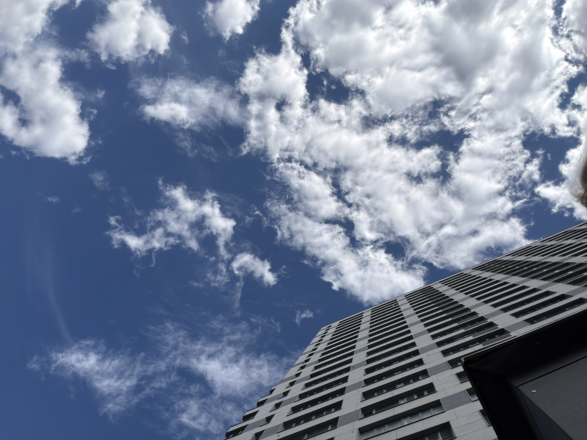 The image captures a low-angle view of a modern, multi-story building against a bright blue sky filled with scattered white clouds. The building's facade is characterized by a grid-like pattern of dark windows. The perspective emphasizes the height and architectural design of the structure. The sky is dynamic, with wispy clouds and some thicker cumulus formations contributing to a sense of depth and movement. The lighting suggests it is daytime, with sunlight illuminating the clouds and casting shadows on the building. The scene appears to be an urban environment, with the building being a prominent feature. There are no visible people or specific landmarks that would definitively place the location within Kazan, Russia, or any other city.