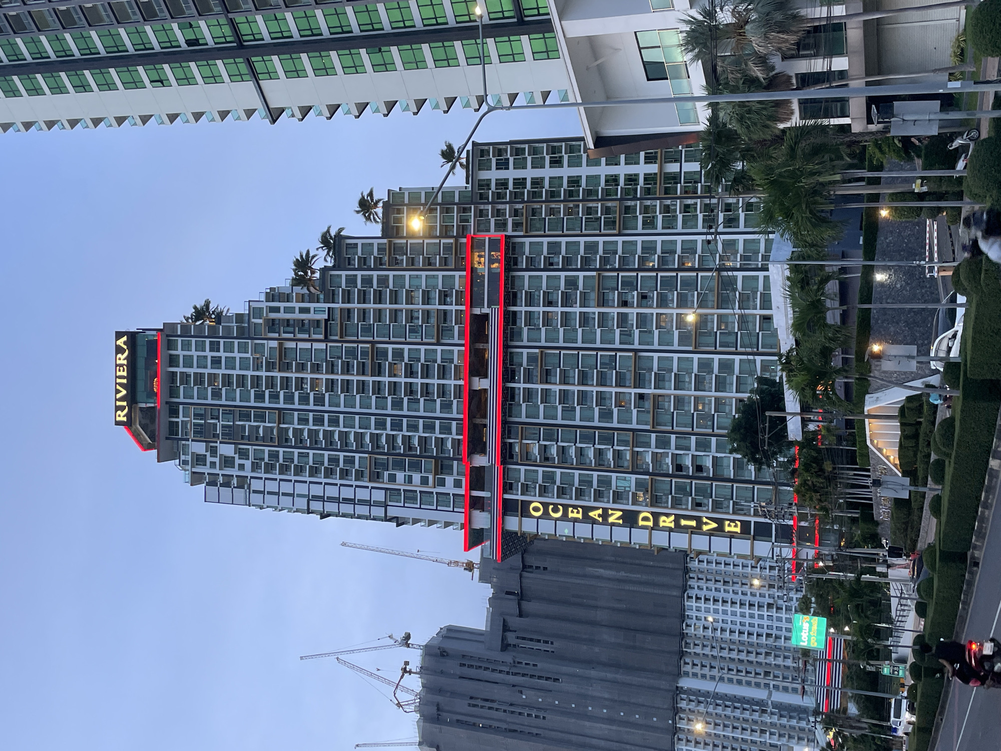 The image captures the Riviera building complex in Pattaya City, Thailand, with "OCEAN DRIVE" prominently displayed vertically in illuminated gold lettering on the facade of one of the towers. A neon red band also runs horizontally across several floors of the building, illuminating several balconies. The top of the building features a sign that reads "RIVIERA". In the background, a tall construction site with cranes is visible, indicating ongoing development in the area. The scene is set against a twilight sky, with streetlights and building lights casting a glow. Palm trees line the street in front of the complex, and several vehicles, including a motorcycle with a rider, are present on the road. Another modern building with green-tinted windows stands to the right of the Riviera complex.