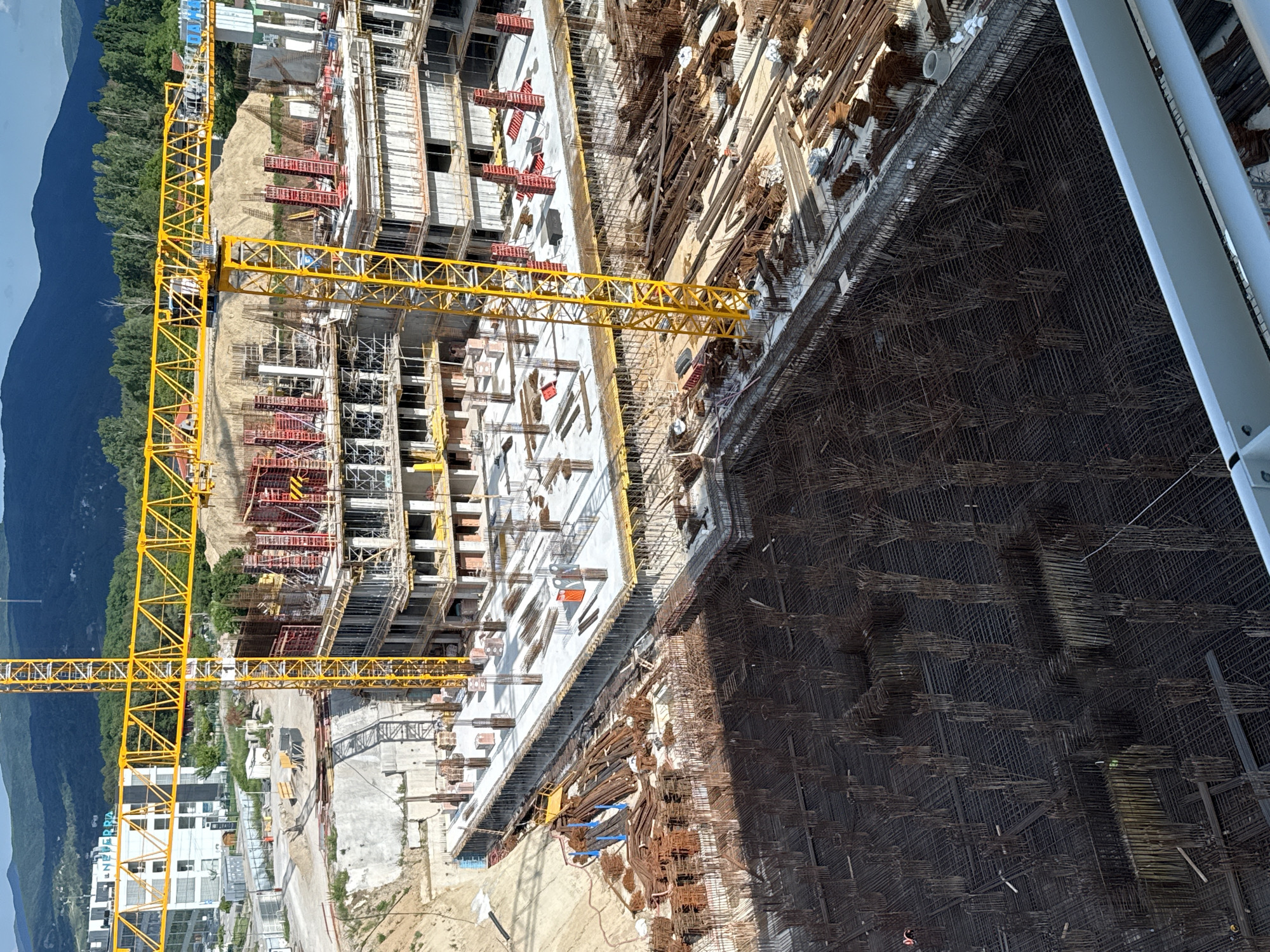 The image depicts a construction site in Sofia, Bulgaria. A large yellow construction crane dominates the left side of the frame. The construction site features several buildings in various stages of completion. Rebar structures are prominent, indicating the ongoing construction of concrete foundations or walls. One building is mostly concrete with some of the rebar structures still showing. In the background, there are mountains, suggesting the construction site is located on the outskirts of the city. A partly completed building is in the bottom left corner, featuring a large sign that says "Le Netterra Park". A large sheet of rebar fencing is directly to the right of the sheet.