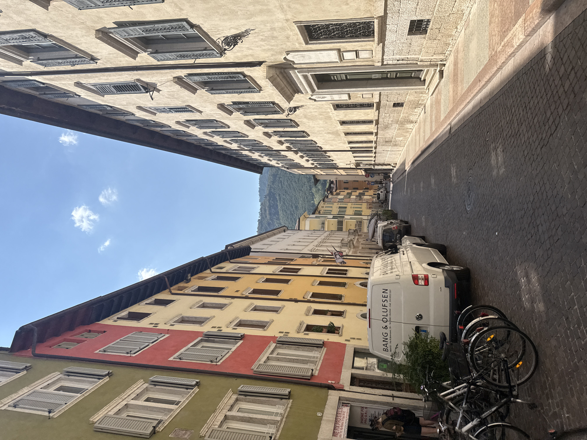 The image depicts a street scene in Trento, Italy. The street is paved with cobblestones, and flanked by tall, colorful buildings on both sides. The buildings have a uniform architectural style, featuring multiple windows with shutters. The buildings on the left are painted in vibrant colors like green, red, and yellow, while the buildings on the right are a lighter, sandy hue.
In the foreground on the left, several bicycles are parked near a white van with the text "BANG & OLUFSEN" on its rear. A person wearing a backpack is standing near the van, looking at their phone. Further down the street, other vehicles and pedestrians are visible.
The sky is clear blue with a few scattered clouds. In the background, a lush green hillside can be seen, indicating the city is situated near mountains. The sunlight casts shadows on the street and buildings, suggesting it is daytime. The overall atmosphere is that of a typical European city street on a sunny day.