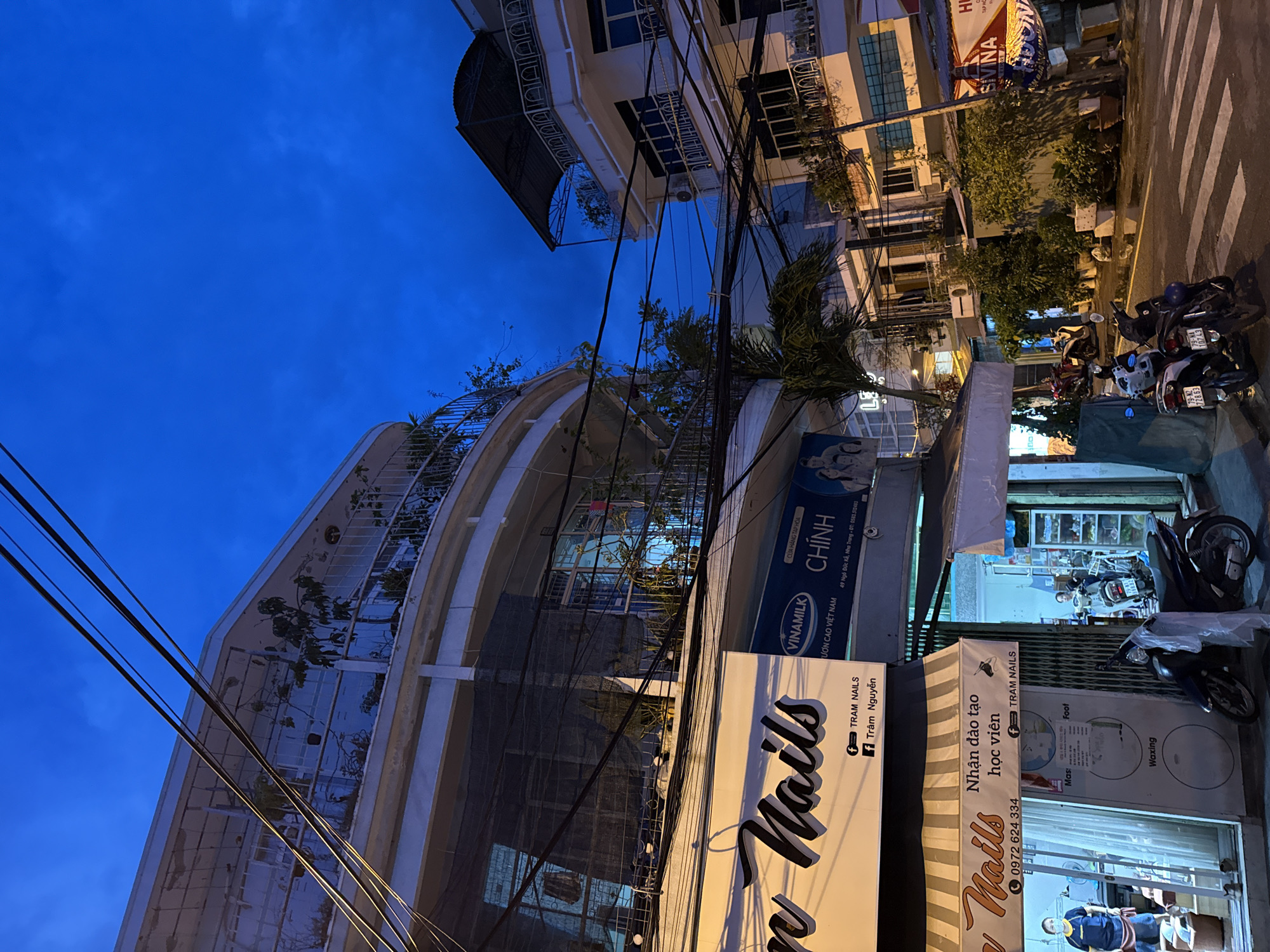 The image depicts a street scene at dusk or night in Nha Trang, Vietnam, with a clear dark blue sky. The foreground is dominated by a street with a pedestrian crossing. To the left, there is a building with a sign that says "Tram Nails" and "Nhận đào tạo học viên," suggesting a nail salon and training center. A person in a blue shirt and mask is visible inside the establishment. There are several motorcycles parked along the street and in front of the businesses.
The architecture of the buildings is characteristic of Southeast Asian cities, with balconies and multiple stories. Wires and cables are prominently strung across the scene, a common sight in urban Vietnam. Further down the street, other businesses and apartment buildings are visible, some with awnings and signage. Greenery in the form of potted plants is present on balconies and near the buildings, adding a touch of nature to the urban environment. The overall atmosphere is that of a bustling city street in the evening, with artificial lights illuminating the storefronts and casting shadows.