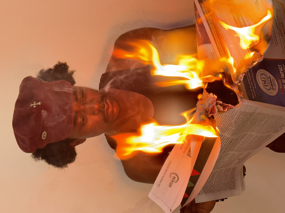 In Igboefon, Nigeria, a dark-skinned individual with dark, curly hair and a mustache is depicted from the chest up, facing forward with a downward gaze. The person wears a maroon flat-top cap featuring a circular emblem on the left side and a gold-colored cross on the front right, along with a dark sleeveless top. Both hands hold an open newspaper in front of their chest, which is actively engulfed in bright orange flames, particularly in the center and right sections. Smoke rises from the burning paper. Visible text on the newspaper includes "BERGER PAINTS NIGERIA PLC" and a blue section advertising "60 YEARS OF IMPACT." The intense fire illuminates the subject's face and upper torso with a strong orange glow. The background is a plain, light-colored wall, also appearing orange due to the fire's light.