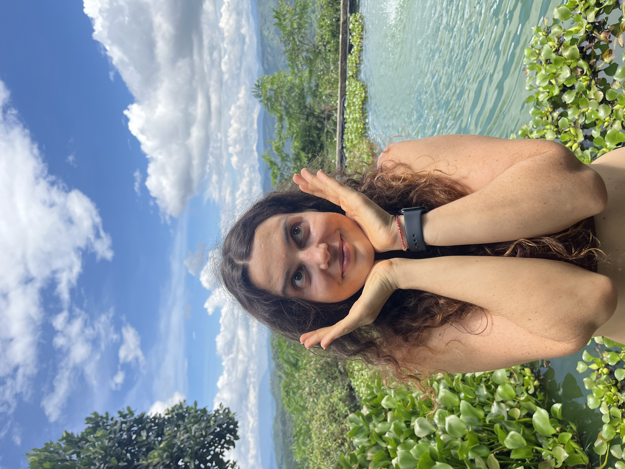 This media file is a portrait of a young woman taken outdoors. She is positioned in the foreground, facing the camera with her head tilted and resting on her hands, which are brought up to her face in a playful gesture. Her hair is long and curly brown, and she is wearing a dark watch on her left wrist. A faint red string is also visible on her wrist. Her expression is a soft smile, and her eyes are looking towards the camera.
The background is a vibrant and lush landscape. To the right of the woman, there is a body of water with some aquatic plants. Beyond the water, a green, hilly terrain stretches into the distance, with trees and vegetation covering the slopes. The sky above is a bright blue with scattered white clouds, suggesting it is daytime and the weather is pleasant. The overall scene evokes a sense of relaxation and enjoyment in a natural setting. The location context indicates this scene is in Sơn Điền, Vietnam. There is no visible text in the media file.