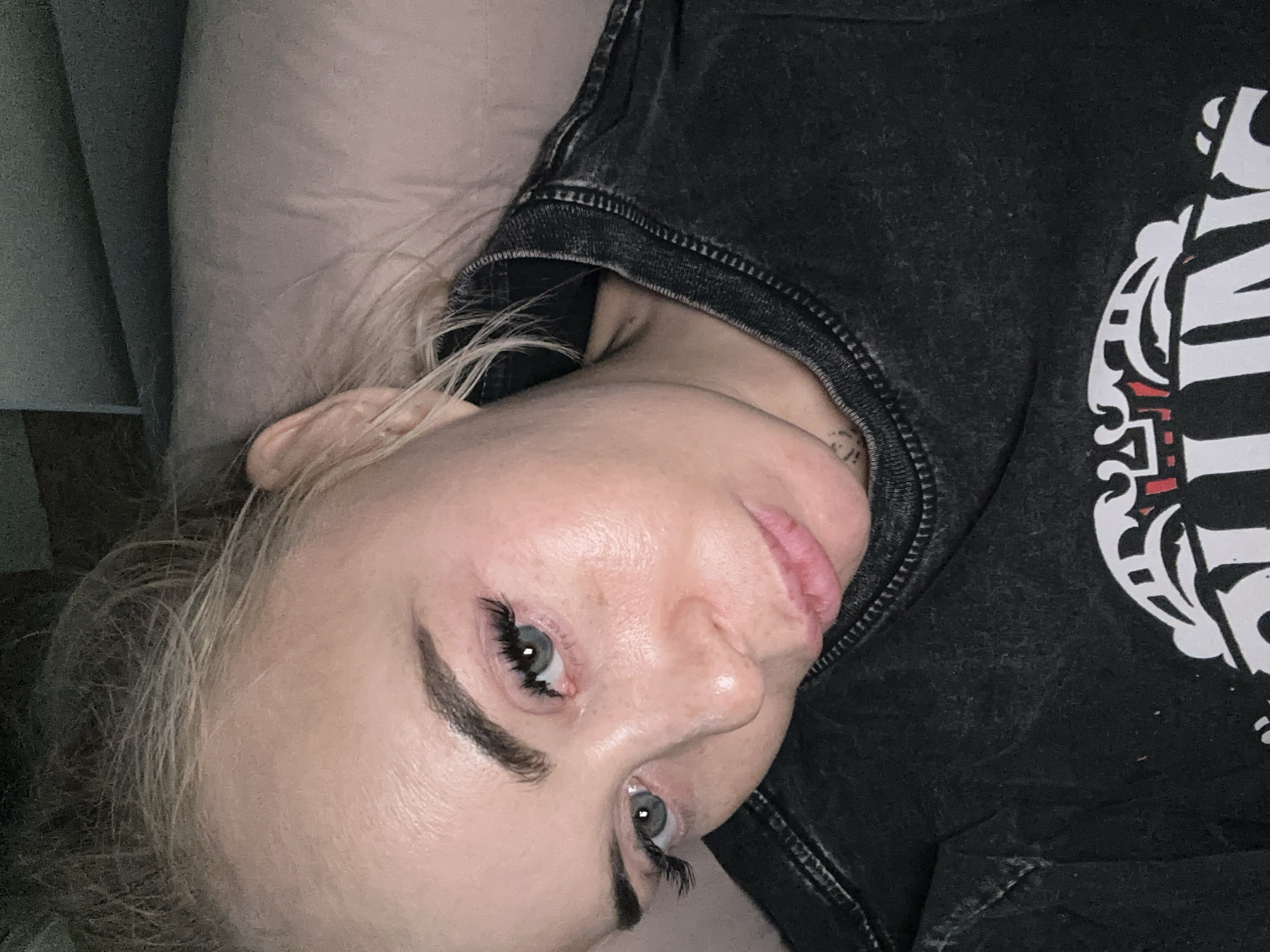 The image shows a person, likely female, with blonde hair, light skin, and striking blue eyes, looking directly at the camera. Her head is tilted to her left, suggesting she is lying down or reclined. She has prominent dark eyebrows and long, dark eyelashes.
She is wearing a dark, possibly charcoal-colored, distressed t-shirt. The shirt features a large white graphic design on the right side, which includes stylized letters, parts of which resemble "IN," along with other decorative elements that could be part of a logo or artistic motif. A small, faint tattoo is partially visible on her neck/shoulder area, beneath the collar of her shirt.
Her blonde hair is visible around her face and shoulder, resting against a light-colored pillow or fabric. The lighting in the scene is dim, indicating an indoor setting, possibly a bedroom, during the evening or night. The person's expression appears neutral or contemplative. This scene is located in Высокий, Russia.