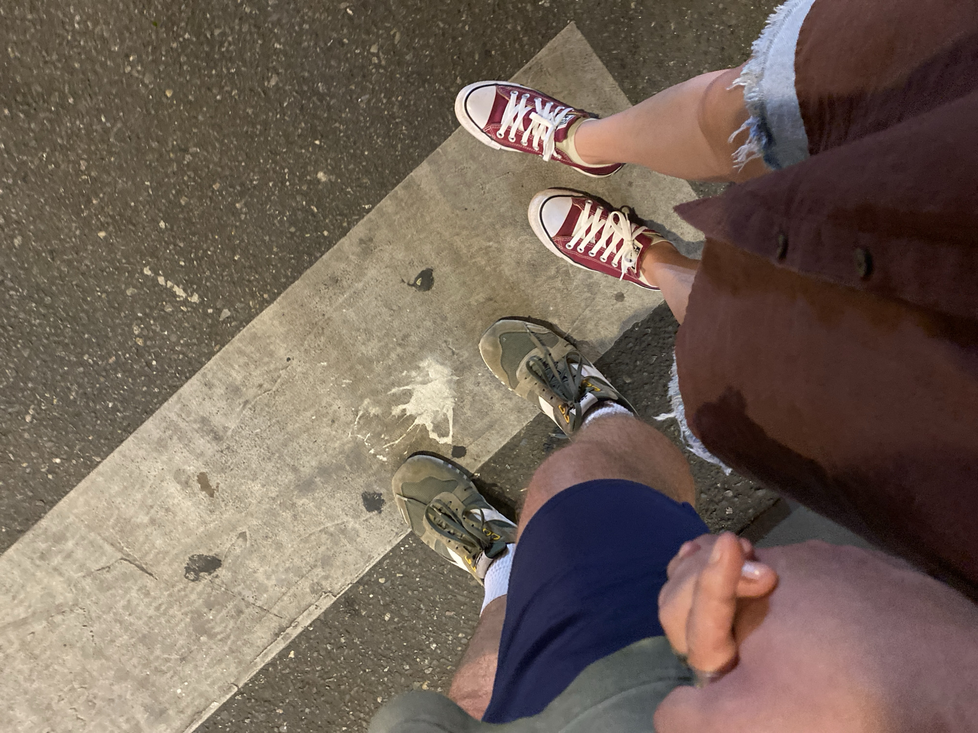 This is a high-angle medium shot of two people holding hands on a street in Batumi, Georgia, at night. The ground is a crosswalk with a grey concrete surface and asphalt. The person on the left is wearing navy blue shorts and green and tan sneakers. The person on the right is wearing a burgundy jacket over denim shorts and burgundy Converse sneakers. Their hands are clasped together in the lower left corner of the frame. There are some white and dark splatters on the crosswalk. The lighting suggests it is nighttime.