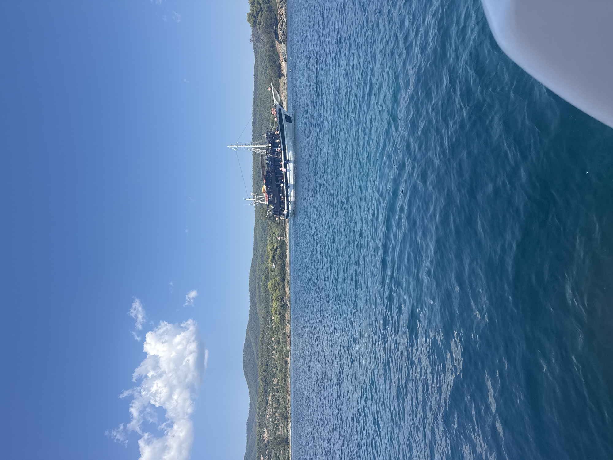 A medium shot captures a clear blue sky with a single fluffy white cloud in the upper left quadrant. Below, a vast expanse of calm, turquoise water fills the majority of the frame. In the mid-ground, a large, dark-hulled boat with white trim and multiple decks is anchored. The boat is populated with several individuals visible on its decks. The background features a lush, green, hilly coastline dotted with trees and vegetation, extending horizontally across the image. The overall scene depicts a serene day at sea, likely in the Sithonia Municipal Unit, Greece, given the characteristic coastline and clear water.