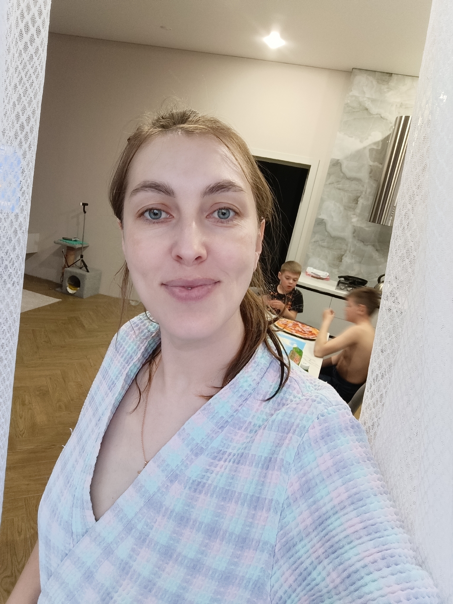 The image, possibly a selfie, shows a woman in a plaid bathrobe, smiling slightly, with two children visible in the background eating pizza at a table. The setting appears to be an apartment interior. In the background, there is a kitchen area with white cabinets and marble-like wall covering. There is also a cat tree in the background. The image was likely taken in Chaikovsky, Russia.
