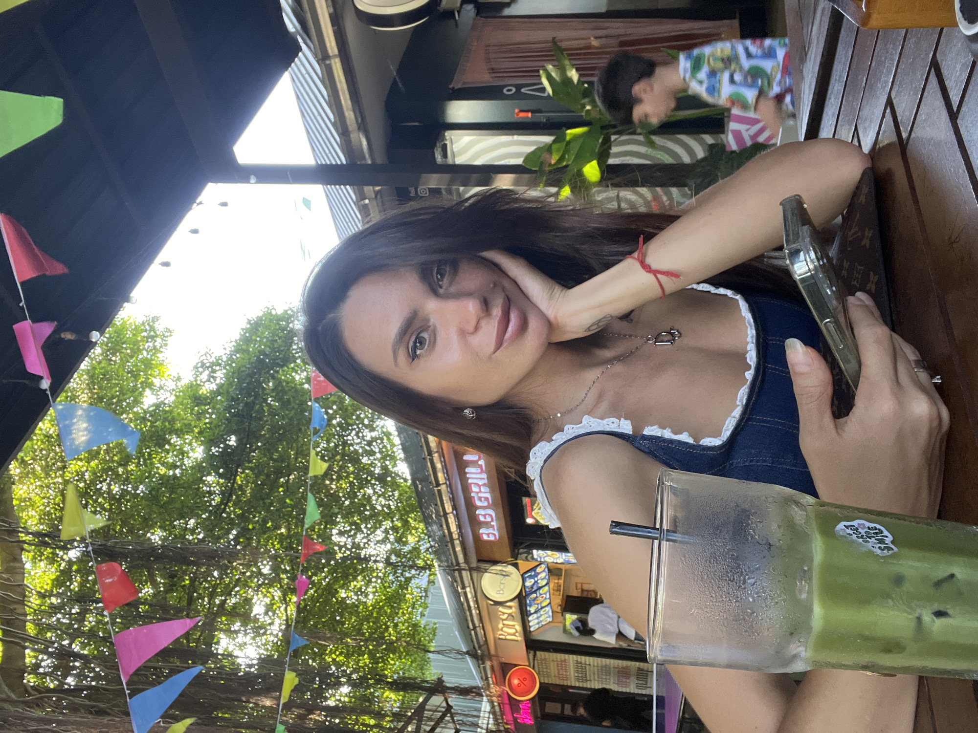 In a bustling outdoor setting with colorful triangular flags strung overhead, a woman with long dark hair and striking makeup is captured in a close-up selfie. She rests her chin on her left hand, adorned with a thin red string bracelet and a silver ring, while her right hand holds a smartphone. A tall glass of green iced beverage with a straw sits on the wooden table in front of her, displaying a sticker with the text "FORGET TO SMILE". Her attire is a denim top with white lace trim.
The background reveals a vibrant street scene, possibly in the area of Ban Raks, Thailand, with awnings providing shade over shopfronts. Signs for "BORSCH GRILL" are visible, and a person in a white cap appears to be working inside one of the establishments. Further back, a young boy in a brightly colored shirt is walking away from the camera. The environment suggests a lively market or dining area, with lush green foliage from overhanging trees contributing to the atmosphere. The lighting indicates it is daytime, with sunlight filtering through the leaves.
