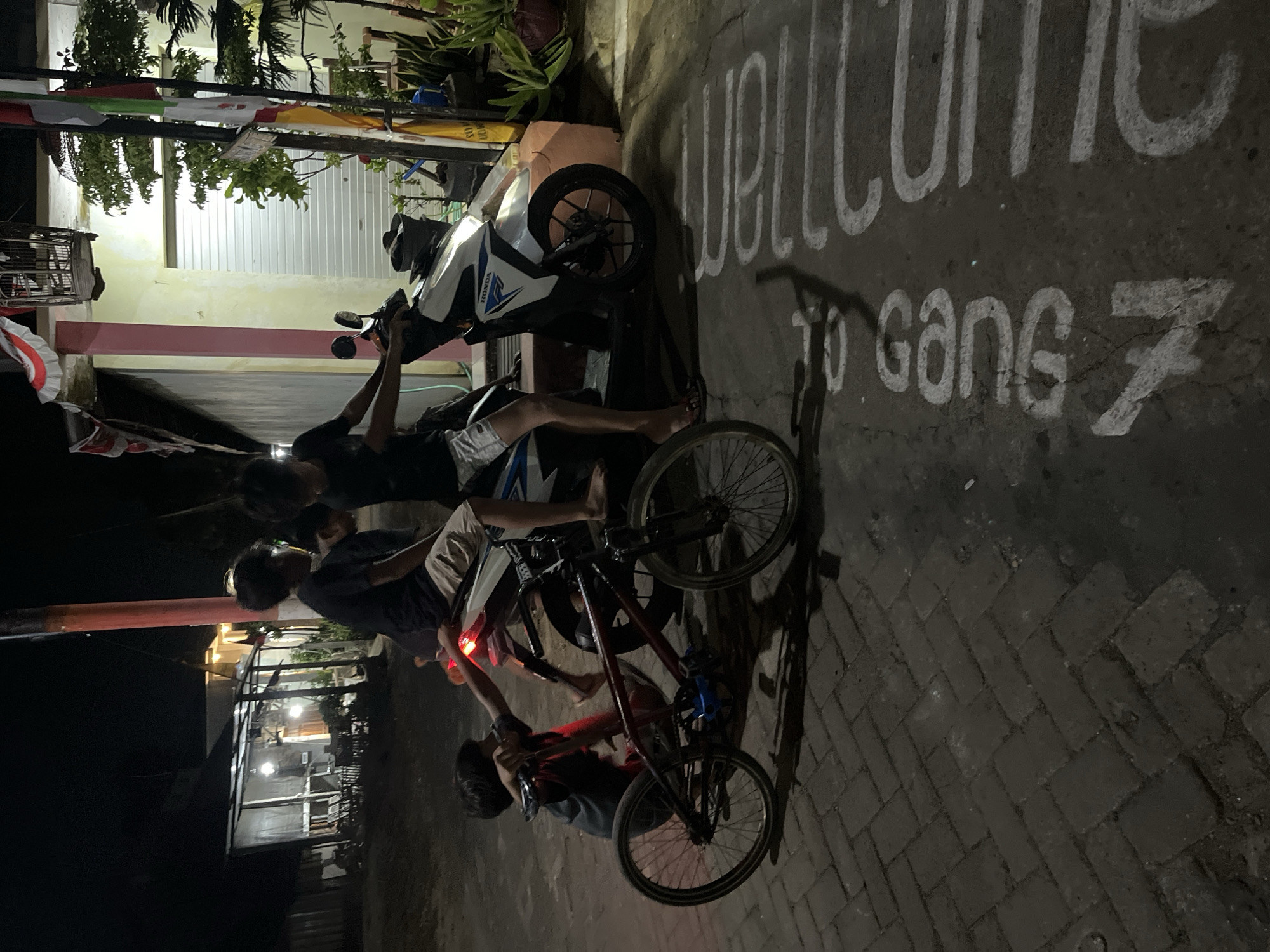 In Karangrandu, Indonesia, at night, a group of young people are gathered around a motorcycle and a bicycle on a paved street. One individual is seated on the motorcycle, holding onto the handlebars, while another person sits behind them. A third individual is actively engaged with a red bicycle, appearing to adjust its handlebars. Another child is positioned between the motorcycle and the bicycle, looking towards the cyclist. The background reveals a building with a light-colored facade and a dark roof, situated in a residential area. The street features a white chalked message that reads "Welcome to Gang 7". The scene depicts a casual nighttime gathering of youth, possibly a street scene in a local community.