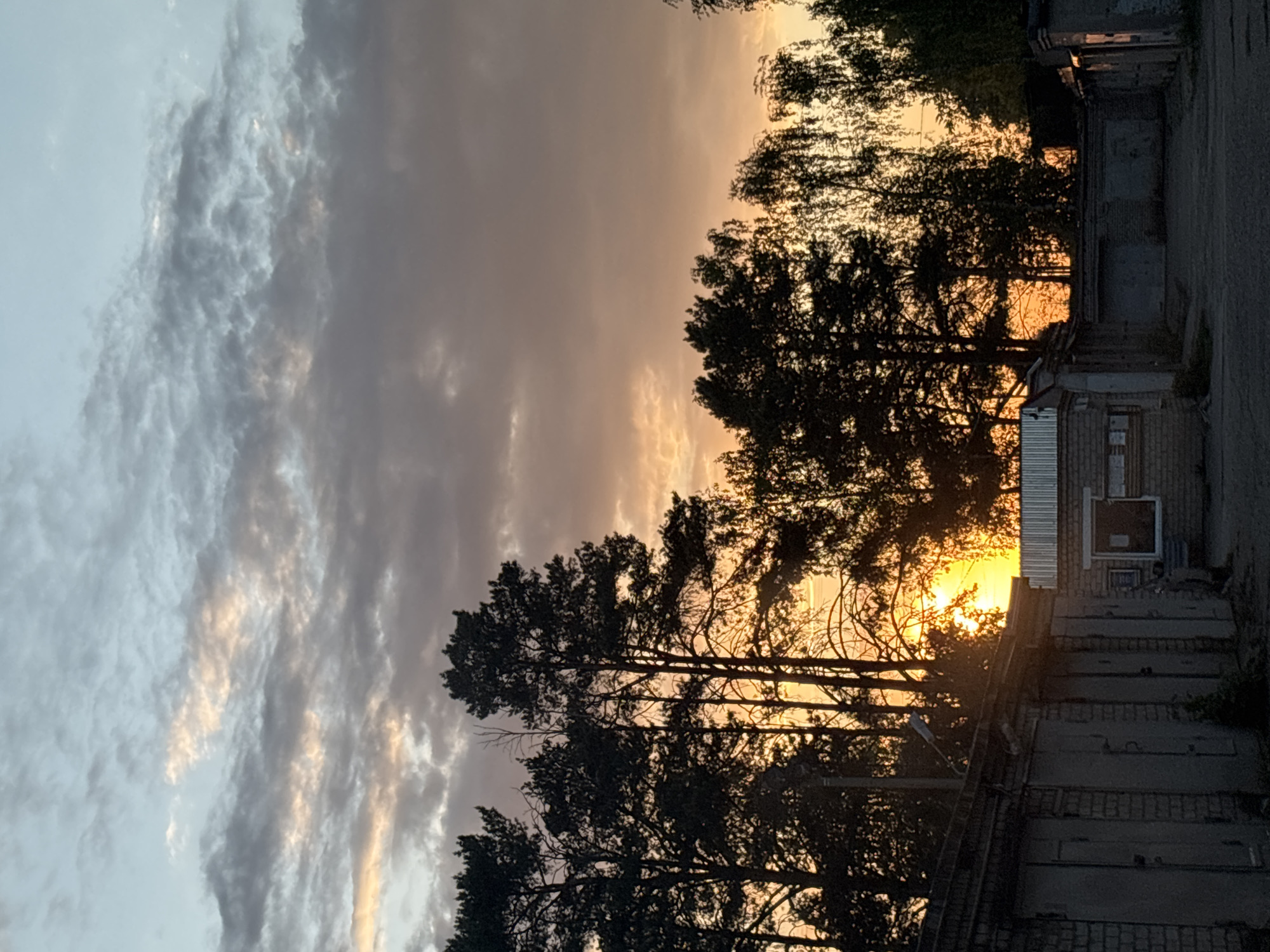 The image depicts a sunset in Chaikovsky, Russia. The sky is a mix of grey clouds and orange light, with the sun appearing to set behind a line of trees. In the foreground, there are a series of garage buildings. The lighting is dim due to the sunset, casting long shadows. There are no people visible in the image.