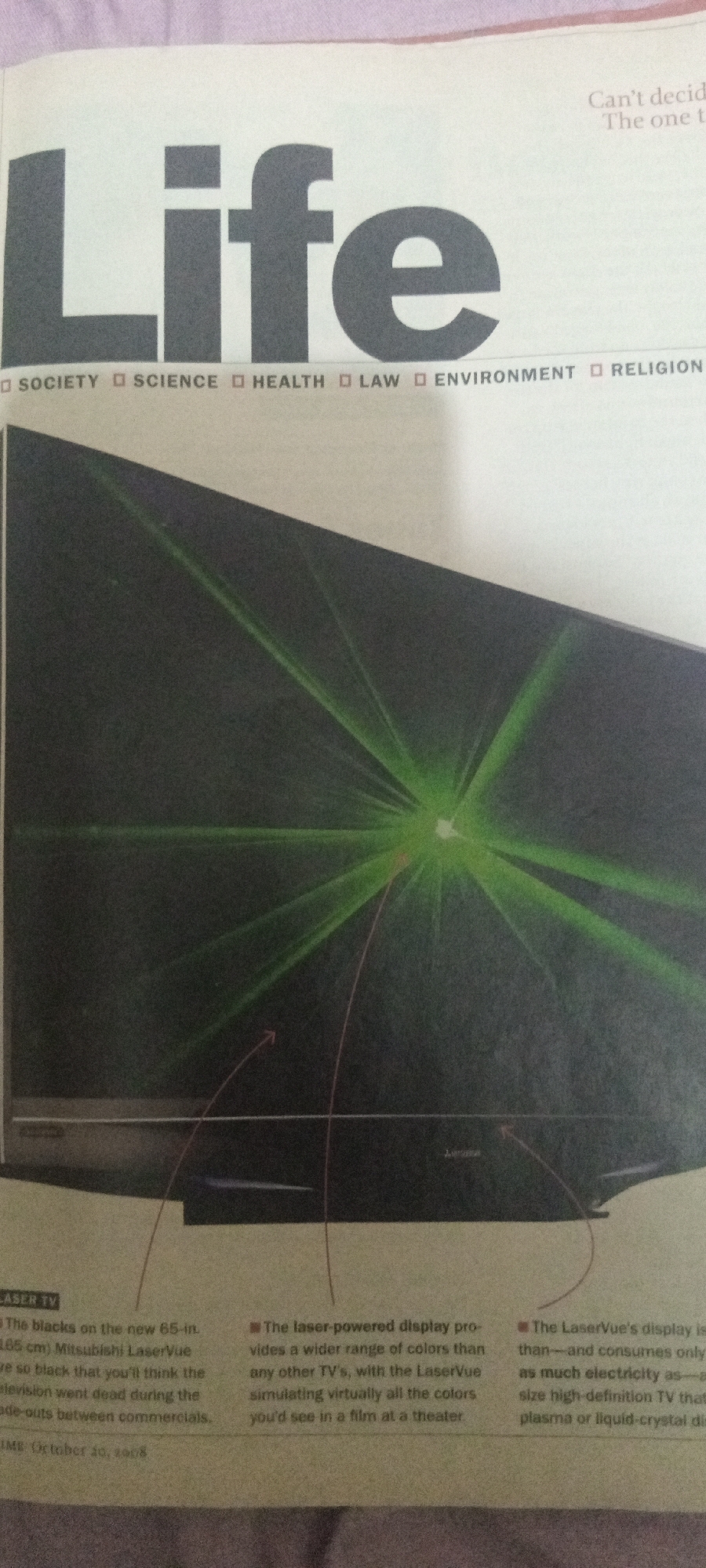 The image shows a magazine cover or a page from a magazine featuring the word "Life" in large, bold, dark gray font. Below the word "Life" are several text entries, including "SOCIETY", "SCIENCE", "HEALTH", "LAW", "ENVIRONMENT", and "RELIGION", each preceded by a small square symbol. To the right of "Life", there is some faint text that appears to be a caption or heading.
The majority of the image is taken up by a photograph of a television screen displaying green laser beams radiating from a central point. The television appears to be a flat-screen model. There are also some red lines and arrows overlaid on the image of the screen, possibly indicating points of interest or demonstrating a concept.
Below the main photograph, there are three text boxes containing descriptions of the television. The first box describes "LASER TV" and mentions a "65-in. (165 cm) Mitsubishi LaserVue" television, noting its black levels and performance during commercials. The second box describes the "laser-powered display" and its wide range of colors, comparing the viewing experience to a theater. The third box discusses the energy efficiency of the LaserVue display compared to other high-definition televisions.
There is no information in the image that indicates its location in the city of Jalingo, Nigeria.
