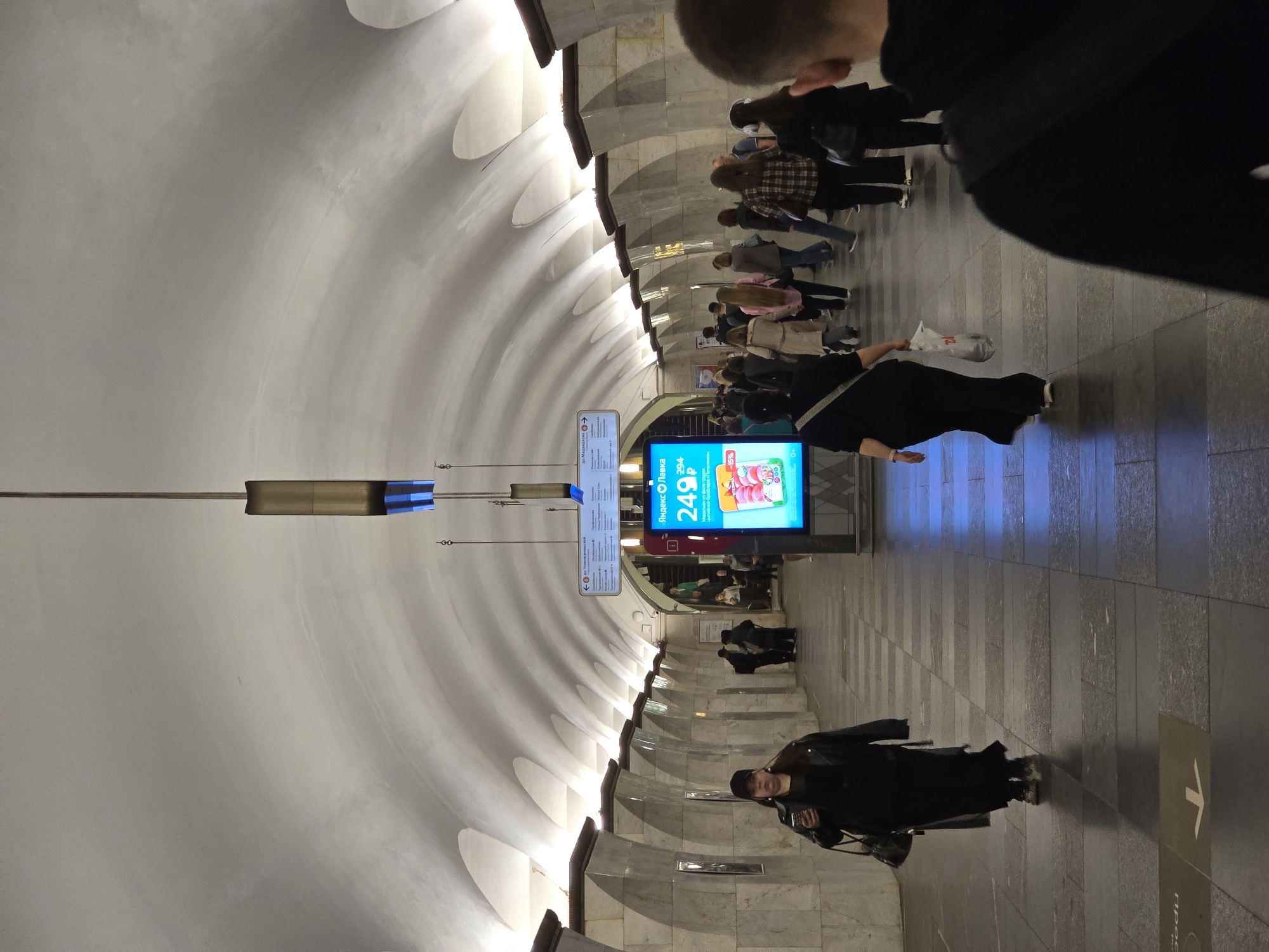 The image depicts the interior of a Moscow Metro station in Russia, possibly Ploshchad Revolyutsii. The station features an arched ceiling and tiled walls and floor. People are moving through the station, some waiting in line. An electronic advertising display is visible.