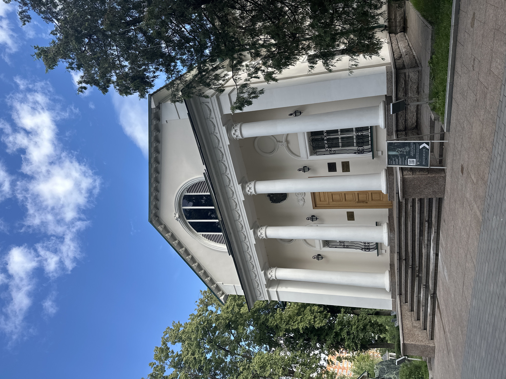 The image shows the exterior of a white classical-style building with large columns and a grand entrance. A semicircular window with black blinds is visible above the entrance. The building is surrounded by lush green trees and set against a bright blue sky with scattered white clouds, suggesting a sunny day. In the foreground, a paved walkway leads up a set of granite stairs to the building's entrance. A sign with an arrow and QR code is placed on the walkway. The scene appears to be a public building, possibly a museum or cultural institution, given the signage. The time of day appears to be daytime. The overall mood is serene and inviting, highlighting the architectural beauty of the building and its natural surroundings. The text on the sign is not clearly legible.