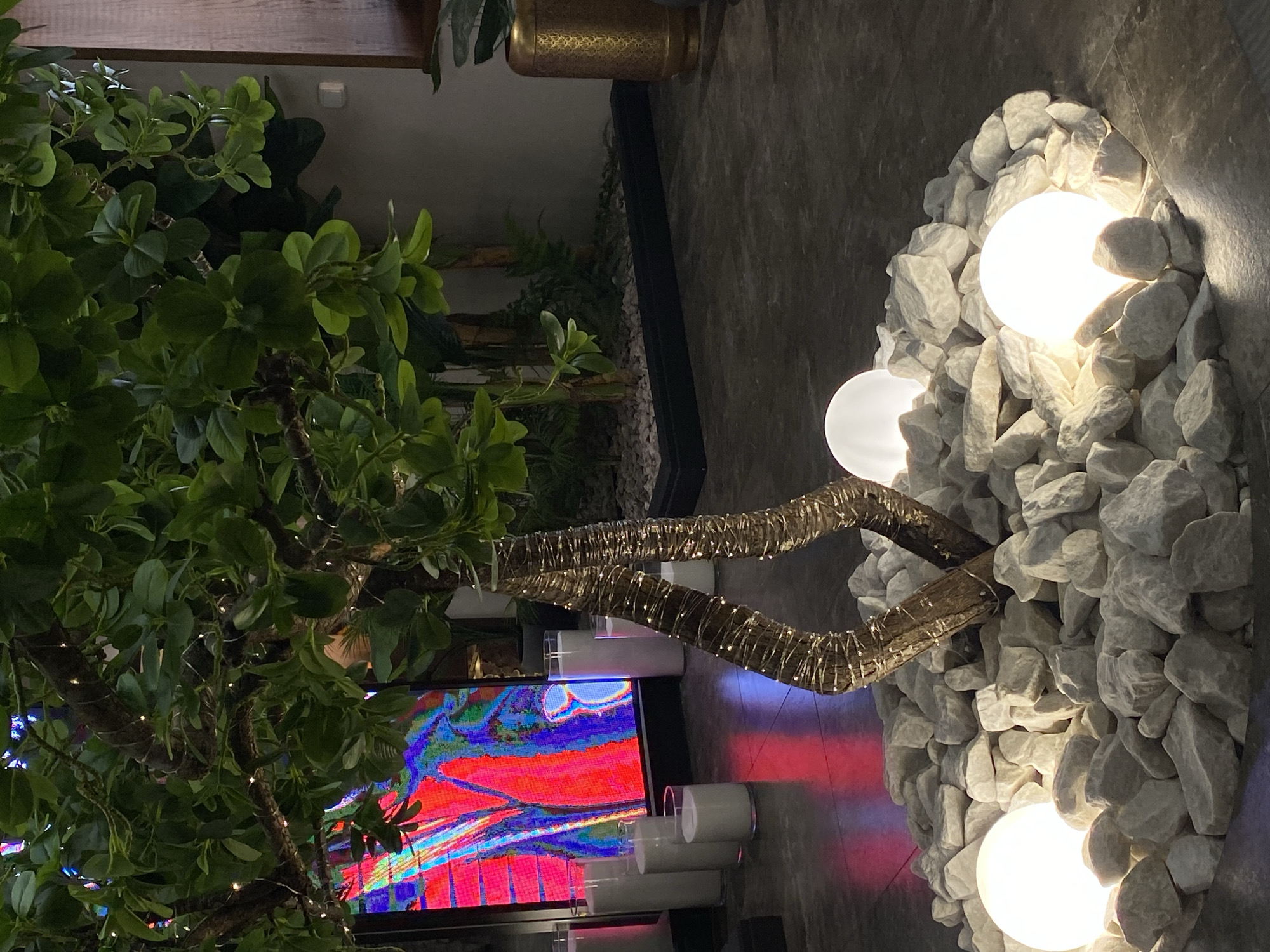 An interior scene in Votkinsk, Russia, displays a decorative plant arrangement illuminated by ambient lighting. Centrally positioned is a substantial artificial tree or plant with numerous green leaves, its intertwining, textured trunks wrapped with delicate string lights. The base of this plant is encircled by a mound of irregular white stones, approximately 15-20 cm in diameter each, within which three illuminated white spherical lights are embedded.
In the background to the left, a large rectangular digital display screen exhibits an abstract image characterized by vivid red, blue, and black pixelated patterns. In front of this screen, several white cylindrical candles, varying in height and housed within clear glass holders, are arranged on the dark floor, casting reflections.
To the right of the central plant, a segment of a golden, ornately patterned cylindrical planter is visible, containing additional foliage. The floor surface is dark and reflective, possibly polished concrete or large dark tiles. The overall environment appears to be an interior space, devoid of human presence, focusing on modern decorative elements and atmospheric lighting.