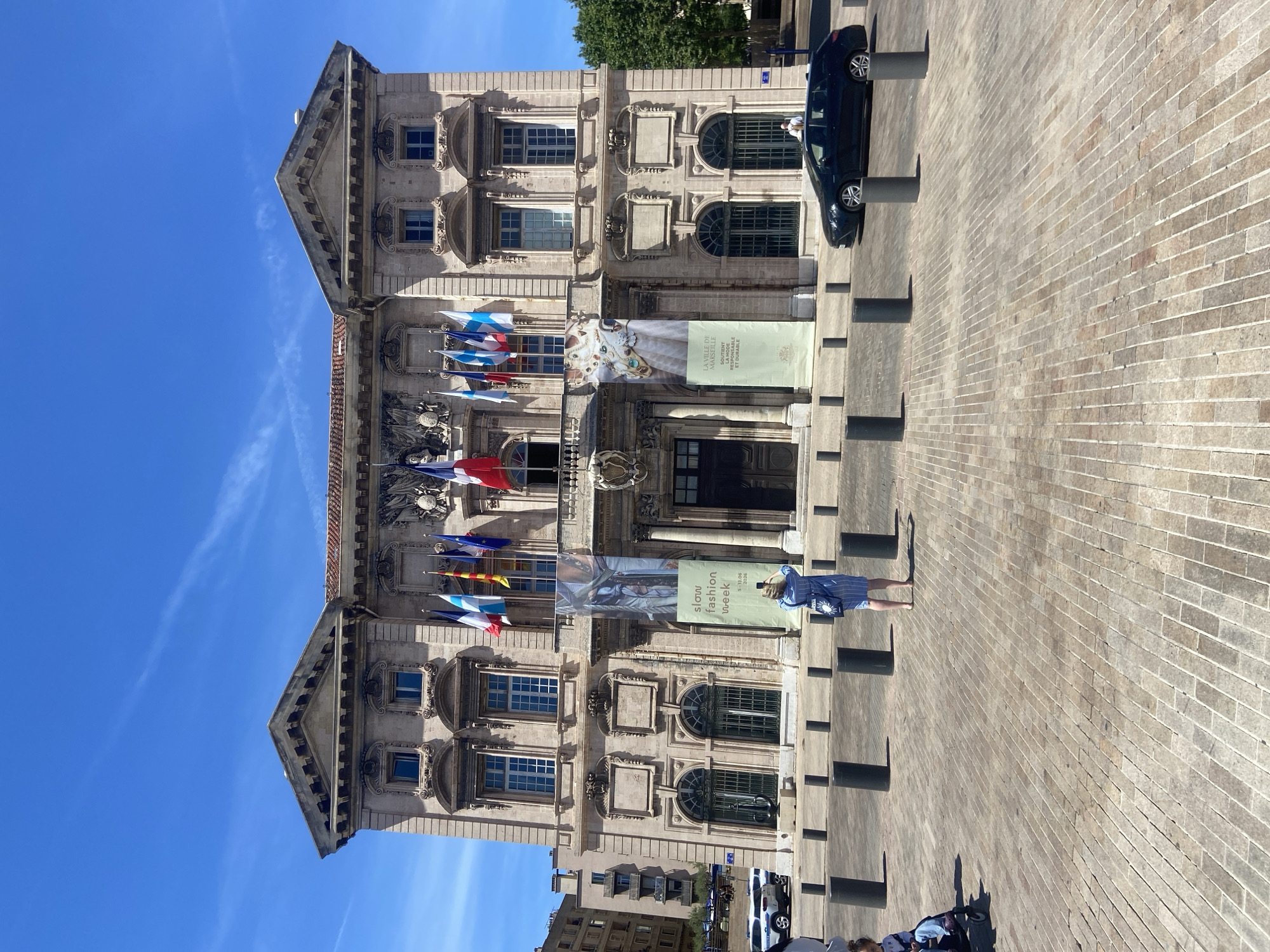 In a bright, daytime scene, the facade of a large, ornate building stands prominently under a clear blue sky with wispy clouds. The building, likely a public institution or government office, features a classical architectural style with multiple stories, arched windows, decorative carvings, and a prominent central entrance. Flags of various nations fly from poles along the upper level of the building. Two large banners hang from the facade: one on the left advertises "slow fashion week" with dates, and another on the right, with text in French, promotes "LA VILLE DE MARSEILLE SOUTIENT UN MODE DE VIE RESPONSABLE ET DURABLE" (The City of Marseille Supports a Responsible and Sustainable Lifestyle).
In the foreground, a paved square is visible, made of rectangular bricks. Several dark, cylindrical bollards are strategically placed throughout the square. A woman in a blue and white striped dress stands in the middle ground, facing away from the camera, holding a smartphone and appearing to take a photograph of the building. To the left, a child in a stroller is partially visible, being pushed by an unseen person. A dark colored station wagon is parked on the right side of the square, near the building. In the background to the left, a portion of another building with balconies can be seen, and to the right, a lush green tree provides a splash of color against the architecture. The overall atmosphere is one of urban civic life on a sunny day.
