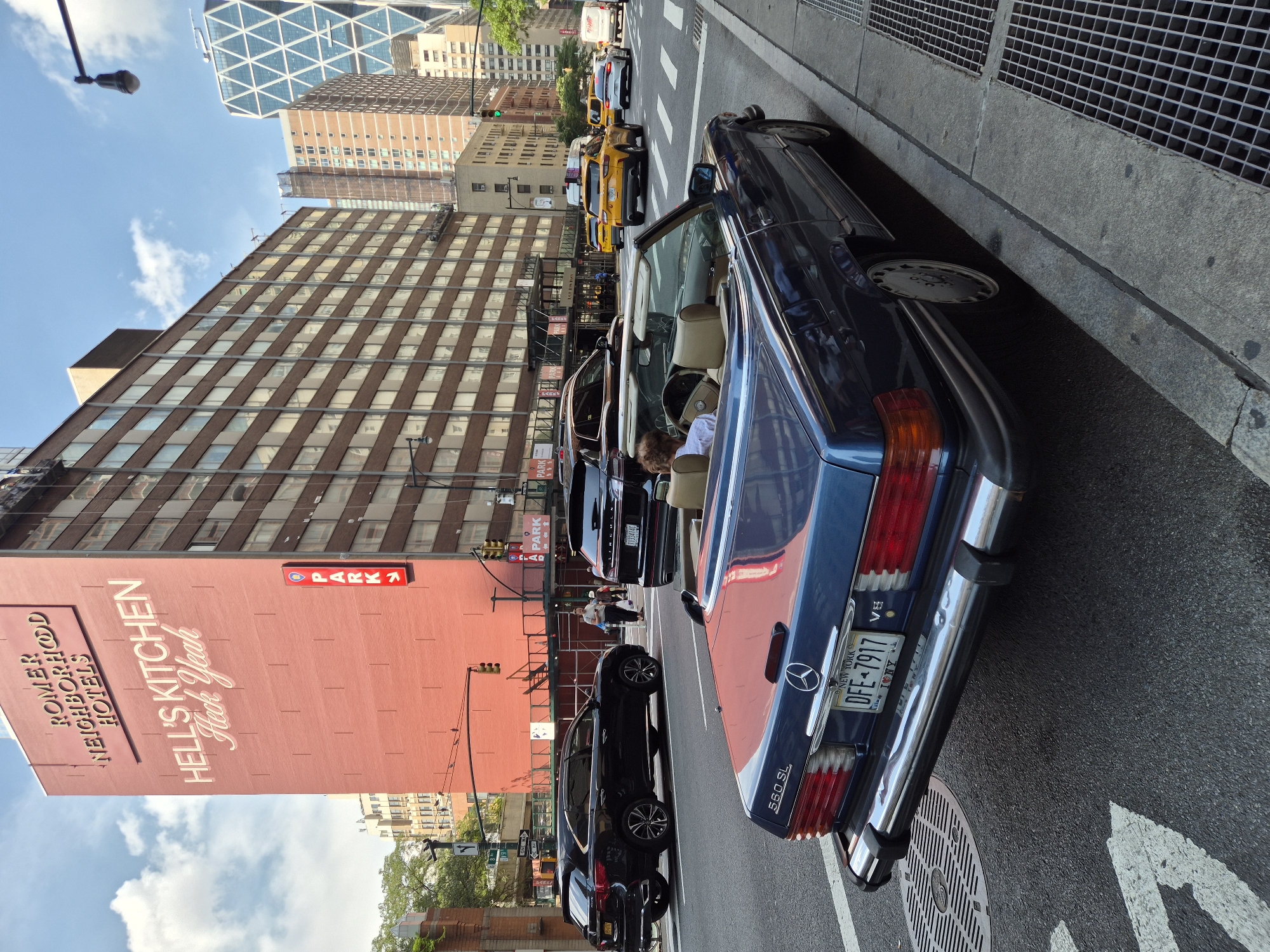 The image depicts a street scene in Hell's Kitchen, Manhattan, New York City, USA. A blue Mercedes-Benz SL convertible is parked at the curb in the foreground, facing away from the camera, with its rear visible. The car's license plate reads "DFE-7917" and displays "NEW YORK" and a heart symbol with "I NY" below it. Inside the convertible, a person with short, light-colored hair is seated in the driver's seat, looking towards the right.
Across the street, a large, red brick building with "ROMER NEIGHBORHOOD HOTELS" and "HELL'S KITCHEN Heck Yeah" painted on its facade is prominent. A vertical red sign with "PARK" and an arrow is attached to the building. Several other vehicles are on the street, including a black SUV and yellow taxis. In the background, various other buildings, including a distinctive modern skyscraper with a diamond-shaped pattern, are visible against a partly cloudy sky. Traffic lights and street signs are also present in the urban environment.