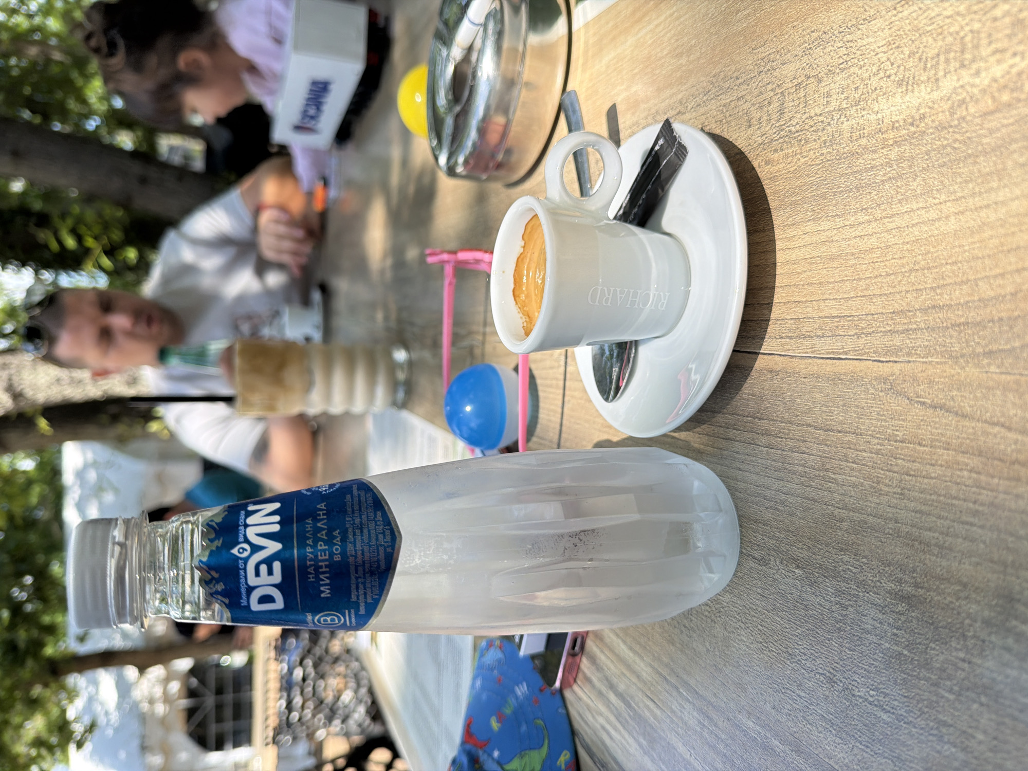 The image captures a moment on a table, likely at an outdoor café or restaurant, given the presence of trees and the sunlit table. The foreground features a bottle of "Devin" mineral water and a white espresso cup from "Richard." The cup is sitting on a white saucer with a packet of sugar. Background includes blurred figures of a man, and a girl playing with a Scania-branded box. The table is a wooden pattern and appears to be outside. The location, suggested by the bottle, is most likely in Bulgaria. Given the urban setting and the presence of other people, it is assumed this takes place in the city of Burgas, Bulgaria.