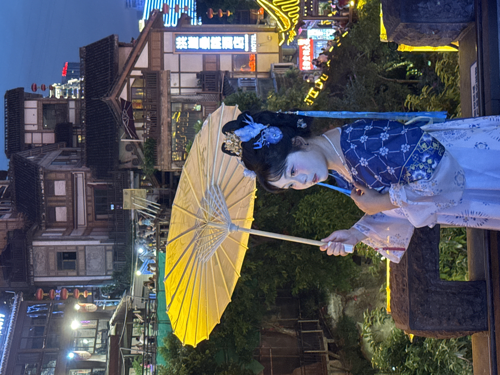 A young woman in traditional Chinese attire, featuring a blue and white patterned robe and an elaborate headdress with blue flowers and tassels, stands on a stone ledge holding a yellow umbrella. She wears a pearl necklace and has makeup on her face. The background depicts a multi-tiered architectural complex with traditional Chinese rooflines and illuminated windows, suggesting a bustling urban environment at dusk or night. Several red lanterns are visible hanging vertically. A neon sign with Chinese characters and the word "HOTEL" is mounted on one of the buildings. The scene is likely located in the Yuzhong District of Chongqing, China, an area known for its distinctive hillside architecture and vibrant nightlife. The woman is posing for a photograph, with her gaze directed to the side and her hand resting on her chest. The overall atmosphere is one of cultural ambiance and urban charm.