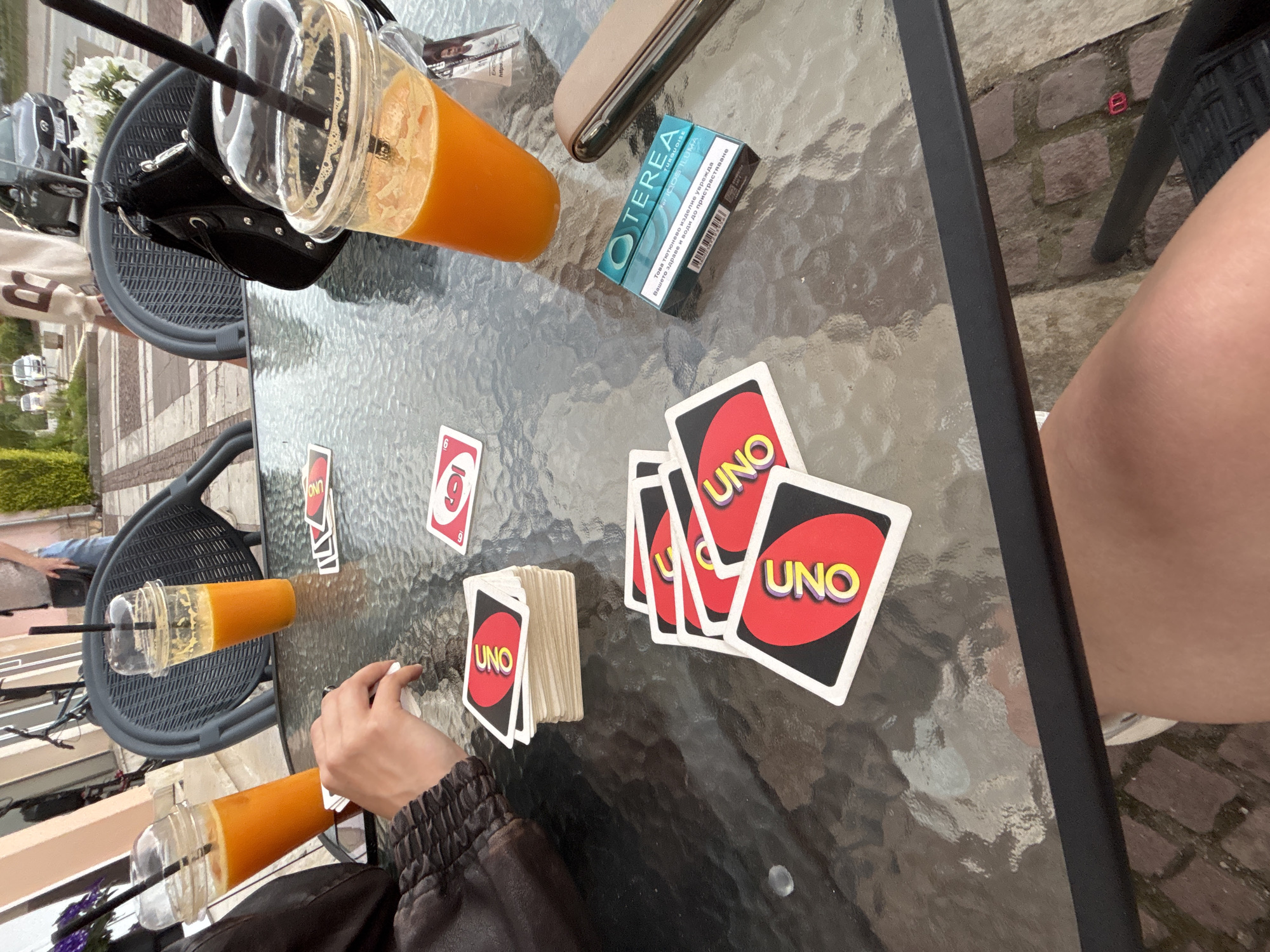 At an outdoor cafe in Garmen, Bulgaria, two clear plastic cups filled with orange juice sit on a glass-top table, each with a black straw. A stack of UNO cards is positioned to the left of the table, and other UNO cards are scattered in front of it. A hand, belonging to an unseen person, is visible reaching for the cards. To the right of the table, a pack of TEREA IQOS ILUMA cigarettes in a teal and blue package sits next to a brown leather case. In the background, people are visible, along with a parked car and a bicycle.
