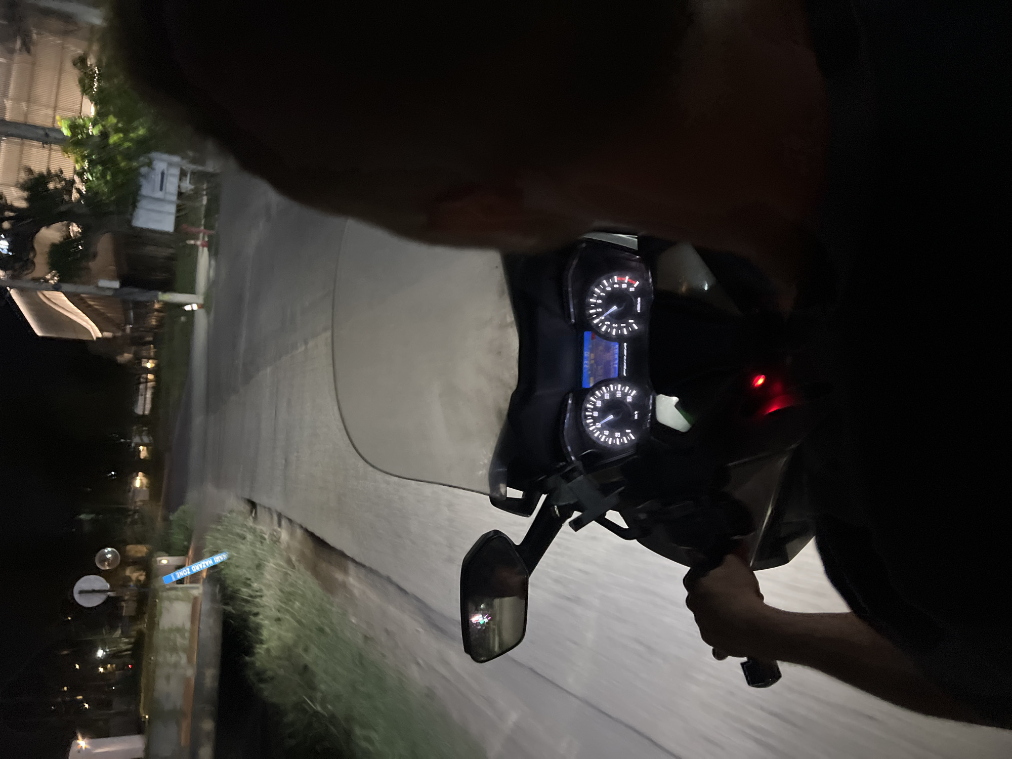 A person is riding a motorcycle on a road at night. The motorcycle's dashboard and side mirror are visible. Street lights and a blue sign that reads "YAH HAZARD ZONE I" are visible on the side of the road.