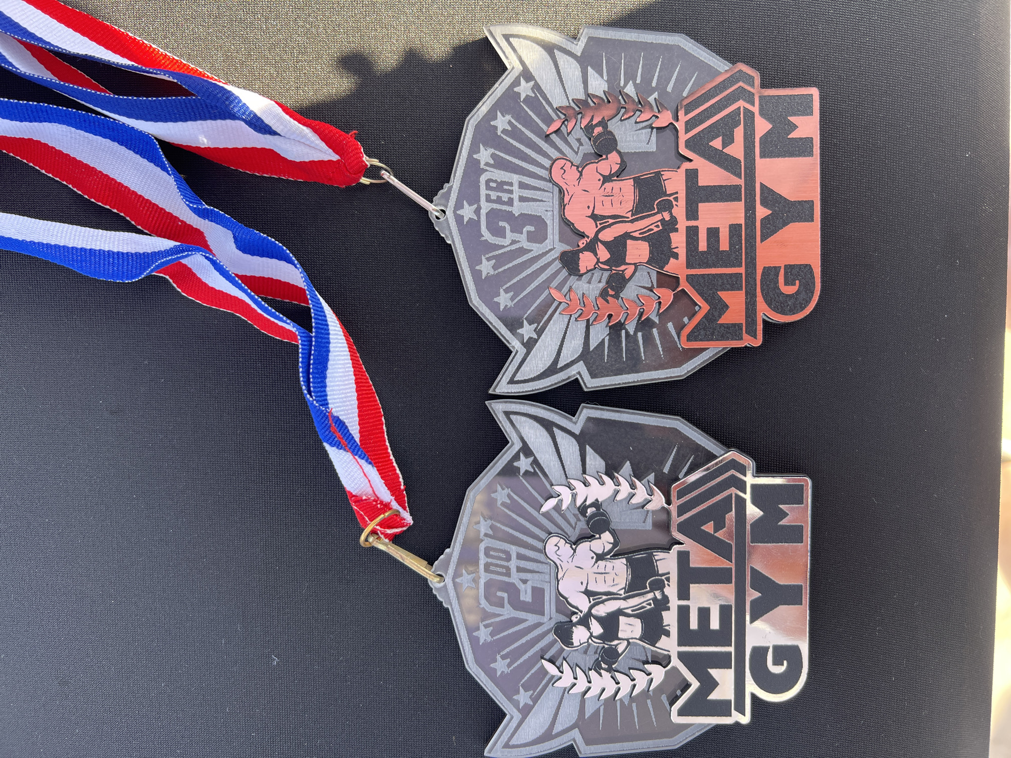 The image shows two medals, one indicating 2nd place and the other 3rd place, both for "Metal Gym." Each medal is attached to a red, white, and blue ribbon. The medals are resting on a dark, possibly black, surface. The setting appears to be outdoors due to the lighting conditions. The location is unknown.