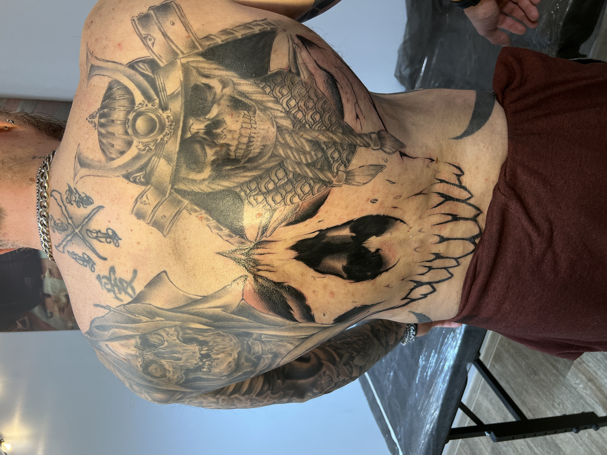 This image captures a person's heavily tattooed back, shoulders, and arms, likely taken in a tattoo studio during or after a session. The subject is a male, identifiable by body hair and a silver chain worn around the neck, and is seen from the back, leaning slightly forward. He wears a dark red or maroon garment pulled down to expose his tattooed torso.
The most prominent tattoo is a large, stylized skull design covering the lower and mid-back, characterized by sharp, jagged edges and significant areas of solid black and grey shading, with subtle hints of red around the eye sockets. On the upper back and right shoulder, another detailed skull is depicted, seemingly armored or helmeted, featuring intricate scale-like textures on a part of the design. An anchor motif is visible on the left shoulder, rendered with detailed shading and ornamentation. Further down on the left arm, more tattoo work includes what appears to be another skull and mountainous landscape elements. Several smaller, indistinct tattoos, possibly script or symbols, are visible on the upper left shoulder and neck area, though no text is clearly legible. The skin around the tattoos shows some redness and slight irritation, suggesting recent tattooing.
In the background, a light-colored wall is visible, and on the right side of the frame, a dark table or trolley covered with clear plastic sheeting, common in tattoo studios for hygiene, is present. A hand, possibly of the tattoo artist, can be seen in one of the crops, reaching towards the subject, further indicating a tattooing process. The flooring appears to be wood or laminate. The overall impression is that of a professional indoor setting, consistent with a tattoo parlor.