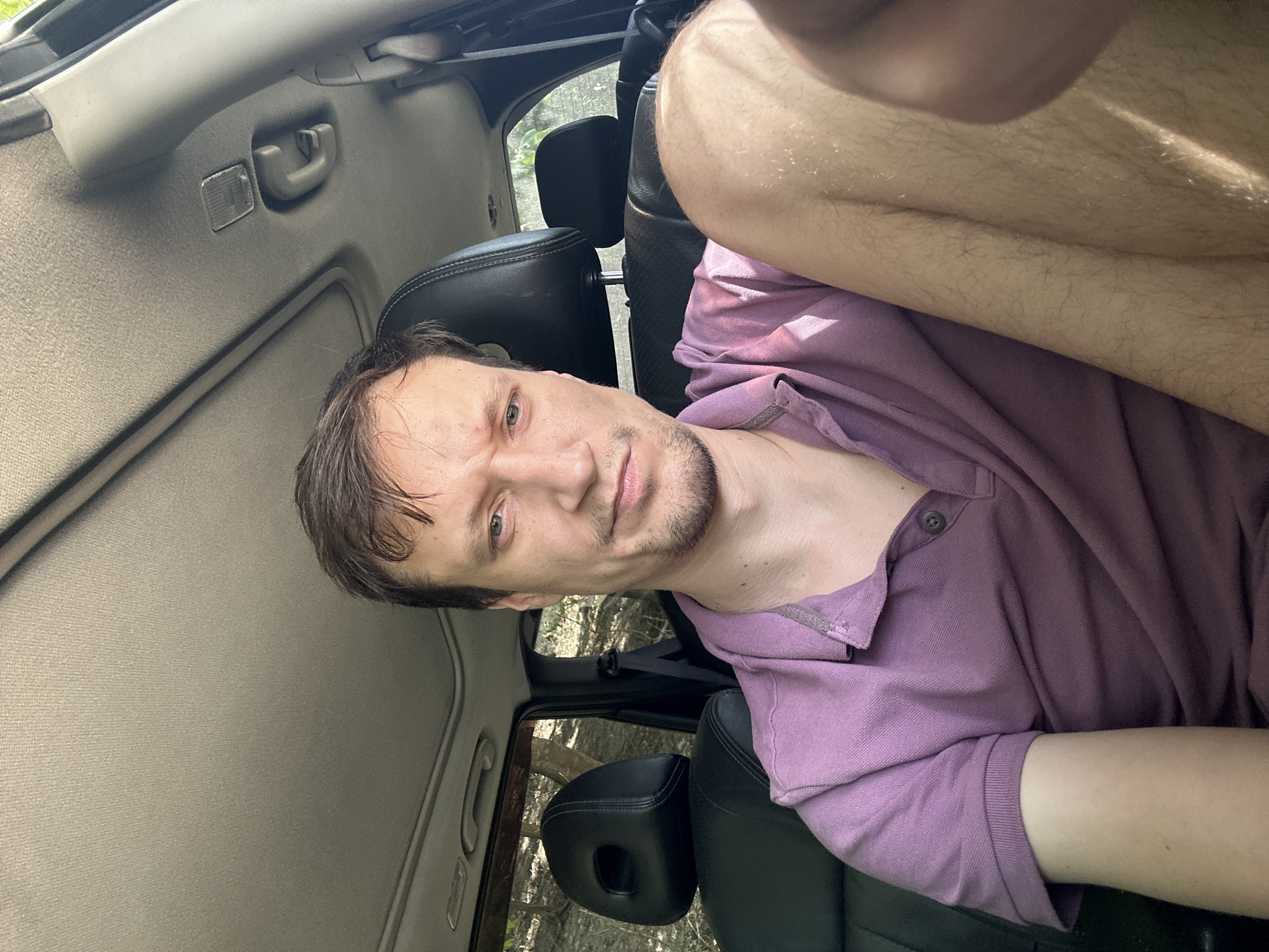 A man with short brown hair and a light beard is seated in the driver's seat of a vehicle, looking at the camera. He is wearing a short-sleeved purple polo shirt. His right arm is bent and rests on his leg. The interior of the car is visible, including the headrests of the seats, the sunroof, and the ceiling of the car. The background outside the car appears to be a wooded area. The image seems to be a selfie taken inside the vehicle, likely in a rural setting given the wooded background. The location of Makiv, Ukraine is not discernible from the image itself.