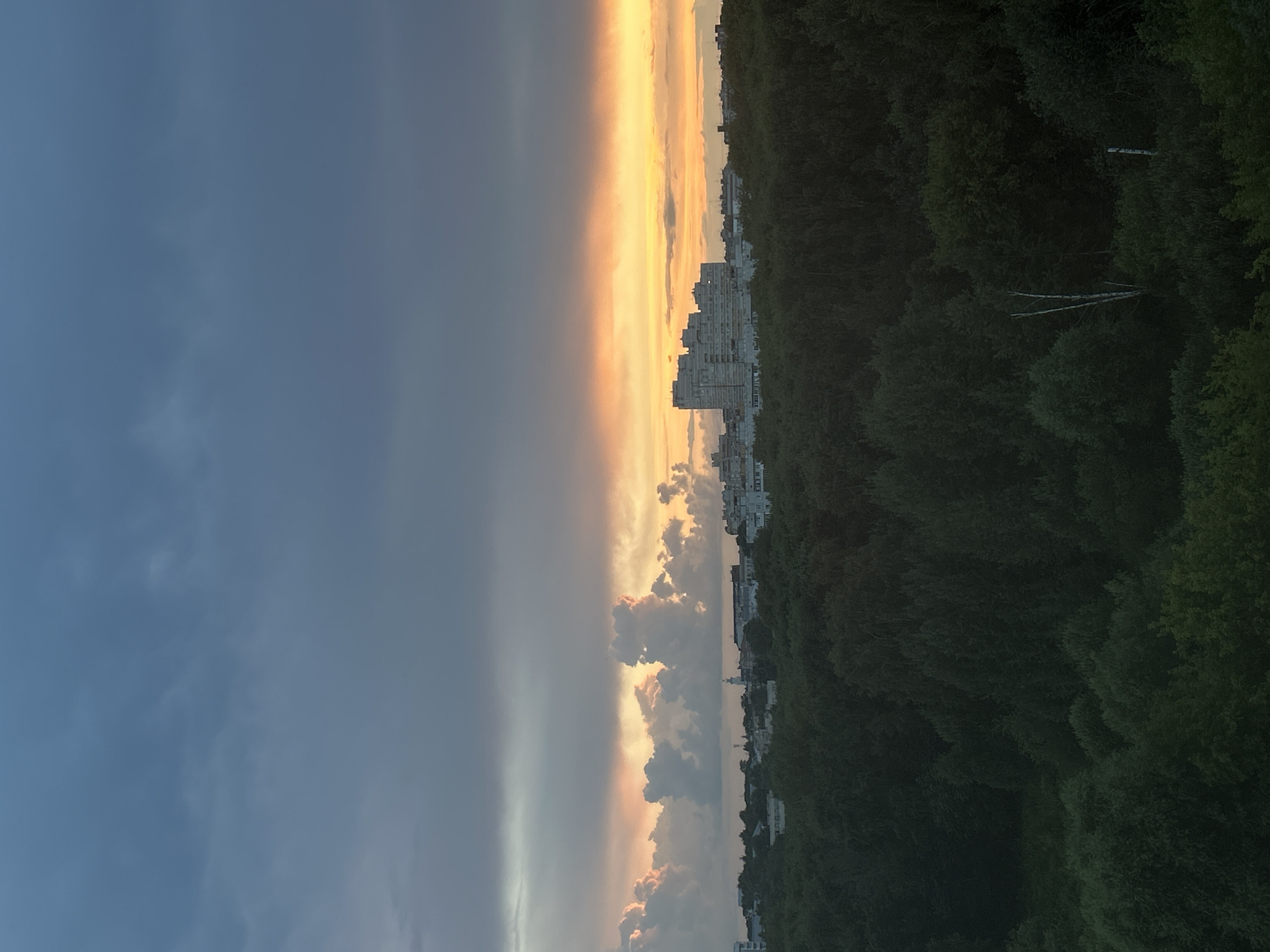 An aerial, slightly high-angle shot captures a dense forest in the foreground, leading to a city skyline in the midground, all beneath a dramatic, clouded sunset. The sky is filled with layers of clouds, with dark gray clouds dominating the upper portion and transitioning to lighter, orange and pink hues near the horizon, suggesting the last moments of daylight. Scattered, puffy white clouds are illuminated by the setting sun. In the distance, the city features a cluster of modern, multi-story buildings, notably a large, light-colored apartment complex in the center, with smaller structures and what appears to be a church steeple visible among them. The forest is lush and dark green, with varied textures indicating different types of trees and foliage. The scene is devoid of any people or active elements.