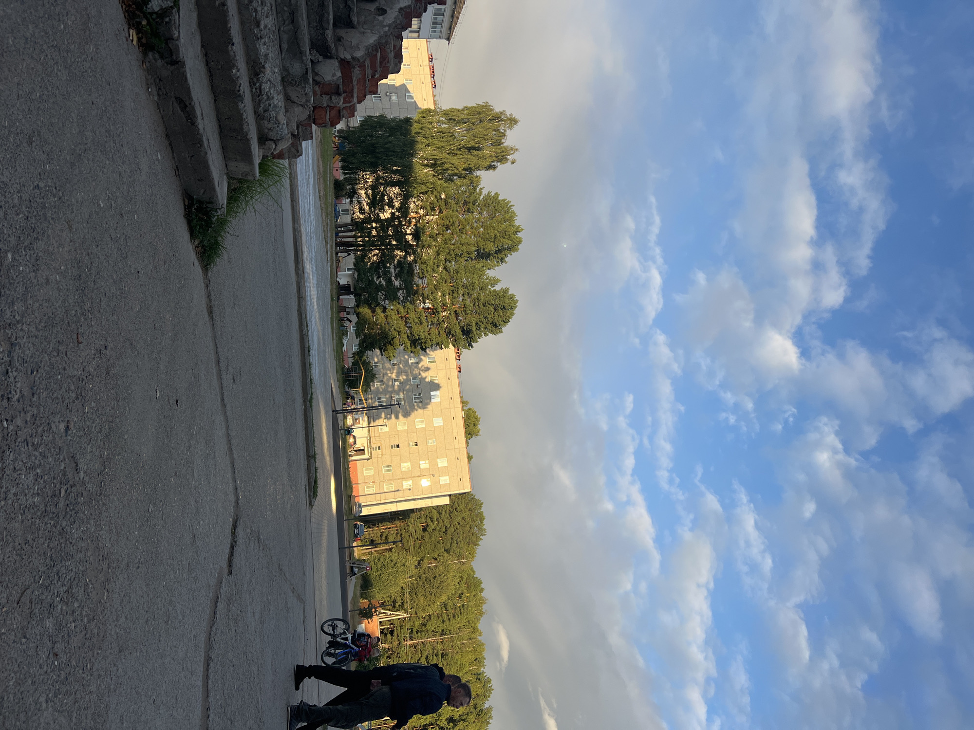 This is a horizontal, eye-level, medium shot of a street scene in Noviy, Russia. In the foreground, there is a set of concrete stairs leading up to the left, made of rough-cut stone and concrete. To the right of the stairs, there is a stretch of cracked asphalt with some sparse grass growing in the cracks. In the midground, there is a paved sidewalk with two men walking away from the camera, one of whom is pushing a bicycle with a child on it. Beyond the sidewalk is a paved street and then a courtyard area with trees and buildings. The buildings are made of light-colored concrete with many windows. Behind the buildings, there is a dense forest. The sky is blue with scattered white clouds, and the sun is setting, casting long shadows.