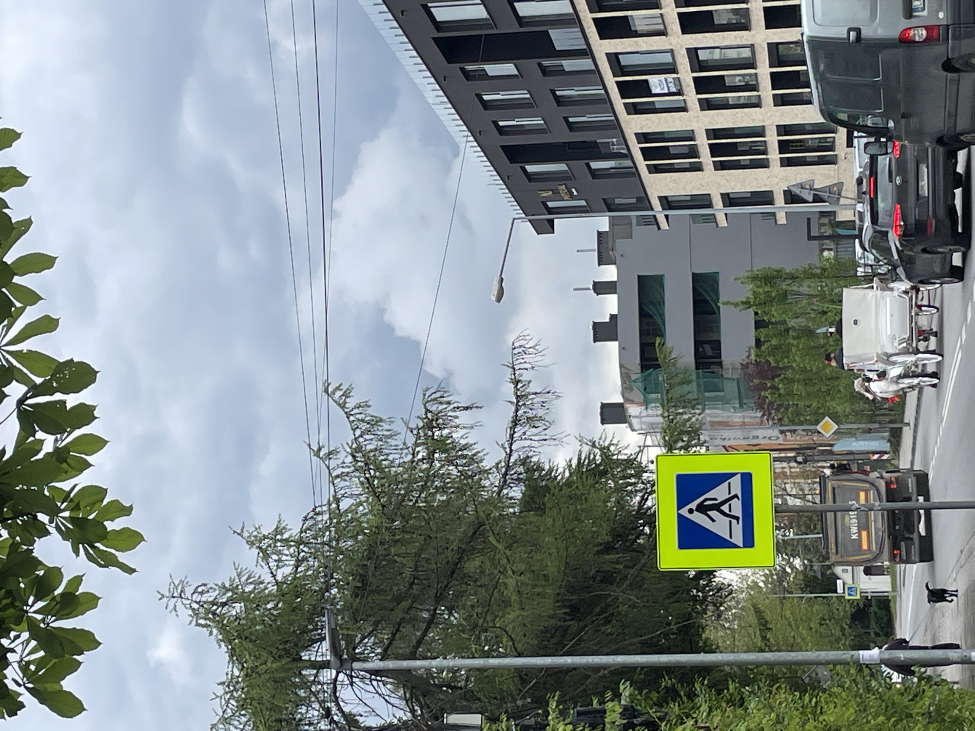The image depicts a street scene in an urban area. Dominating the foreground is a pedestrian crossing sign with a yellow border and a blue square containing a white silhouette of a person walking. In the background, a multi-story building with a modern, geometric facade is visible. Several vehicles, including a gray van and a black car, are lined up on the street. Two individuals dressed in white protective suits appear to be working near the vehicles. Power lines stretch across the sky. The sky is overcast with a layer of clouds. Trees and greenery frame the bottom of the image. A black dog is on a leash on the right side of the frame. A truck with the license plate KW19195 can be seen on the road.