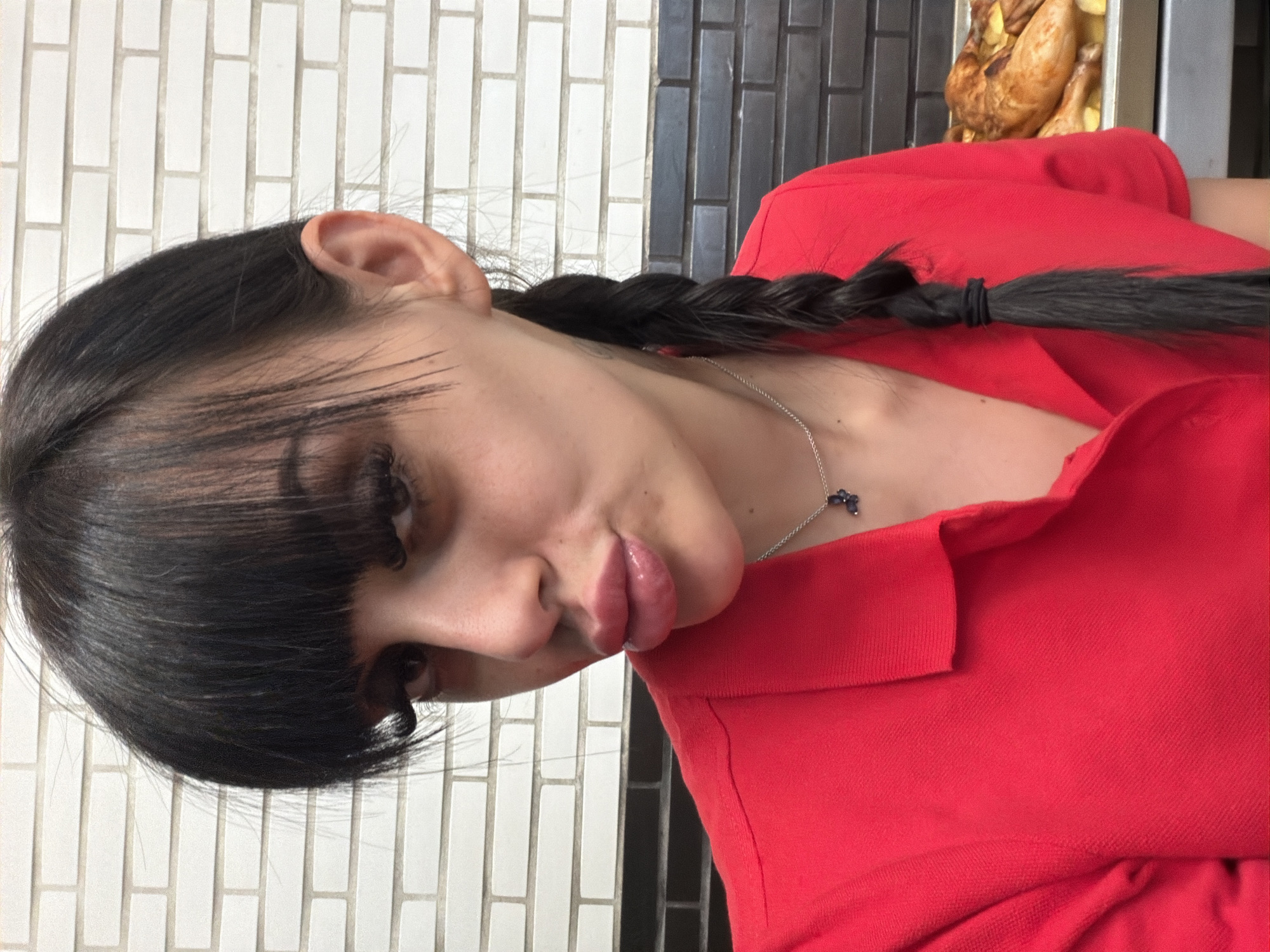 The image shows a close-up of a woman with dark hair styled in bangs and a braid, wearing a red collared shirt and a silver necklace with a small, dark pendant. She is making a kissing face, with her lips pursed. Her left eye is partially obscured by her bangs. In the background, a brick wall is visible, with alternating rows of white and dark grey bricks. To the right of the woman, a tray of cooked chickens is partially visible. The setting appears to be indoors, possibly a restaurant or food service establishment. There are no discernible features that would indicate the location is Plovdiv, Bulgaria.