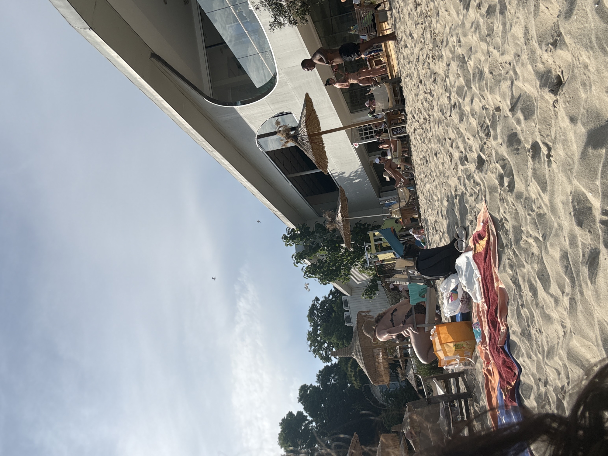 The image depicts an outdoor beach scene during daylight, captured from a low perspective. In the immediate foreground, dark, blurred hair is visible on the bottom left. The lower half of the frame is dominated by light-colored sand with visible undulations and footprints. On the sand, a striped beach towel (red, orange, blue, and white) is laid out, accompanied by a yellow and orange reusable shopping bag, a white plastic bag, a black backpack, and a pair of white sandals or flip-flops.
In the mid-ground, a person with long, light-colored hair, wearing a patterned bikini top, is seated on a wooden chair under a straw umbrella (palapa). This person is facing away from the camera, looking towards the left. To their left, another wooden chair is partially visible. Behind them, lush green trees and foliage form a dense backdrop, with additional straw umbrellas scattered amongst them.
To the right, a distinctive modern building with a light-colored facade dominates the background. The building features a large, rectangular upper level with dark-tinted windows reflecting the sky. A prominent, large circular cut-out detail is integrated into the building's facade on the right side, also reflecting the sky. Below the upper level, an outdoor patio or deck area is visible, furnished with tables, chairs, and several straw umbrellas.
On the sandy area in front of this patio, a shirtless man with dark skin, wearing dark swim shorts and sunglasses, stands facing left, holding what appears to be a mobile phone or small object. Behind him, another person in a light-colored bikini is partially visible. Further left on the patio, individuals are seated at tables, one person notably with their legs propped up.
The sky is a pale blue with scattered white clouds. Three birds are visible flying: one in the lower right quadrant of the sky, and two smaller ones higher up towards the left of the building. The scene is brightly lit by natural sunlight, casting subtle shadows on the sand and objects.
