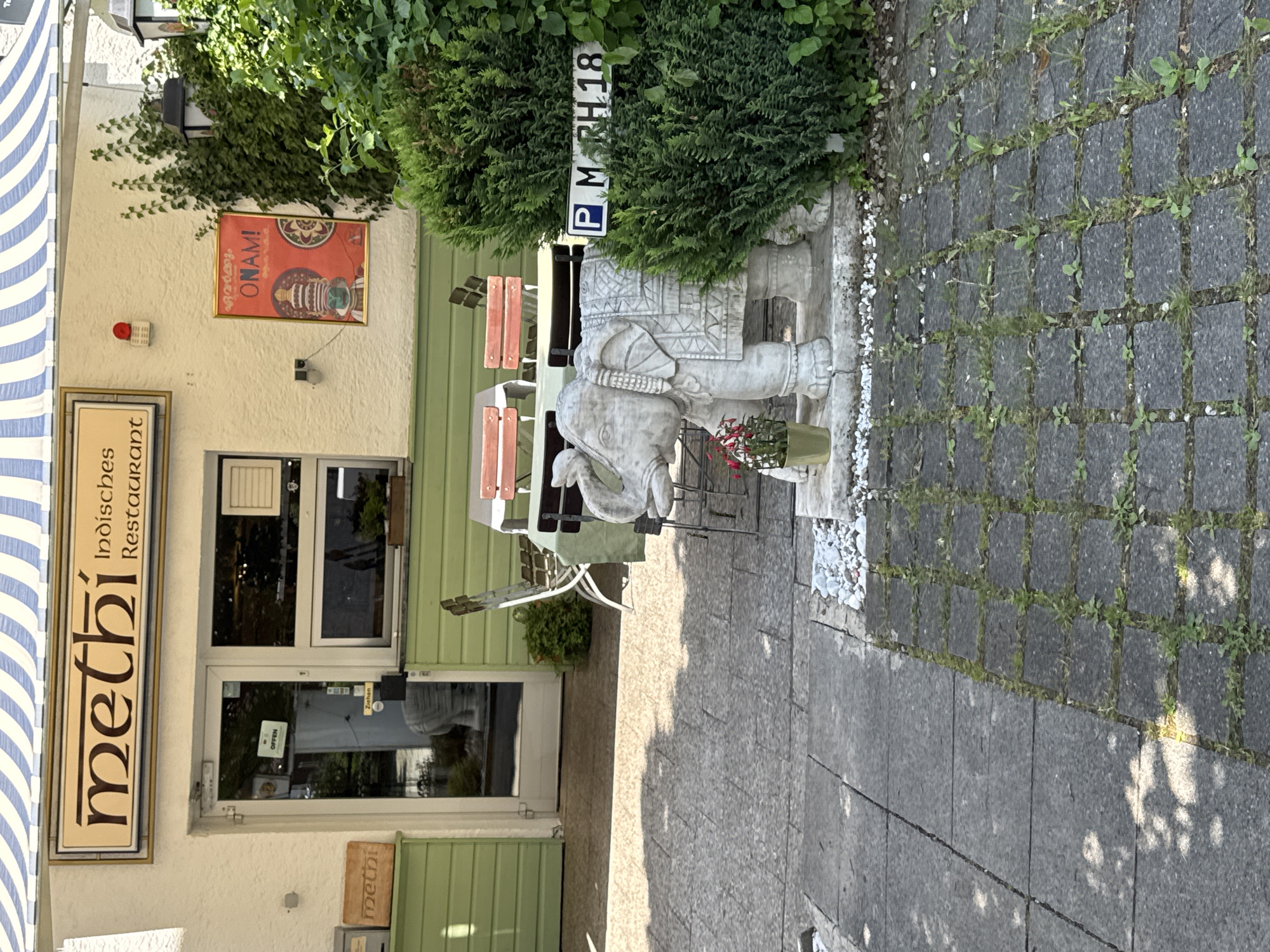 The image shows the exterior of an Indian restaurant named "methi Indische Restaurant" in Höhenkirchen-Siegertsbrunn, Germany. The restaurant has a light-colored facade with a blue and white striped awning over the entrance. A sign above the entrance reads "methi Indische Restaurant". To the left of the entrance, there is a small wooden sign with "methi" written on it. The entrance is a glass door with a sign inside indicating "OFFEN" (Open). Adjacent to the door, there's a window and a green colored wall.
In front of the restaurant, an outdoor seating area is visible with a table and chairs. A prominent feature in the foreground is a stone statue of an elephant, which is intricately carved and decorated. Next to the elephant statue, there is a small potted plant with red flowers. A green bush is located to the right of the elephant, and a parking sign with "P MRH 18" is attached to it. The ground is paved with stone tiles, some of which have grass growing in the gaps. Sunlight casts shadows on the pavement. A poster with the word "ONAM!" and an illustration of a traditional Indian figure is displayed on the wall to the right of the entrance.