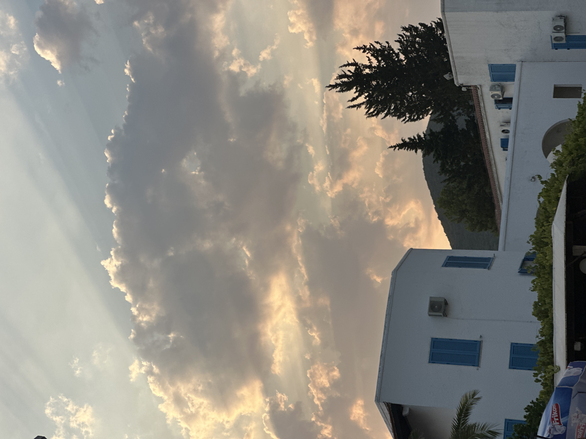 The image depicts a serene evening scene in Budva, Montenegro. The sky is filled with a dramatic display of clouds, illuminated by the warm hues of sunset. Sunbeams pierce through the clouds, creating a soft, ethereal glow. In the foreground, two white buildings with distinct blue shutters are visible. These buildings appear to be part of a hotel or residential complex, common in coastal towns. Lush green foliage surrounds the buildings, hinting at a well-maintained outdoor area. Tall, dark green cypress trees stand prominently against the backdrop of a rolling, verdant hill, likely a characteristic of the Montenegrin landscape. The overall impression is one of tranquility and natural beauty.