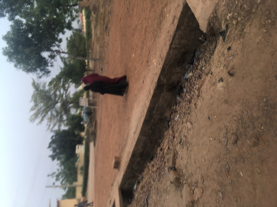 A woman in a red and black robe walks along a dirt road. The road is flanked by trees and shrubs on one side and a concrete drainage ditch filled with debris on the other. The sky is overcast. A person can be seen walking in the distance on the left.