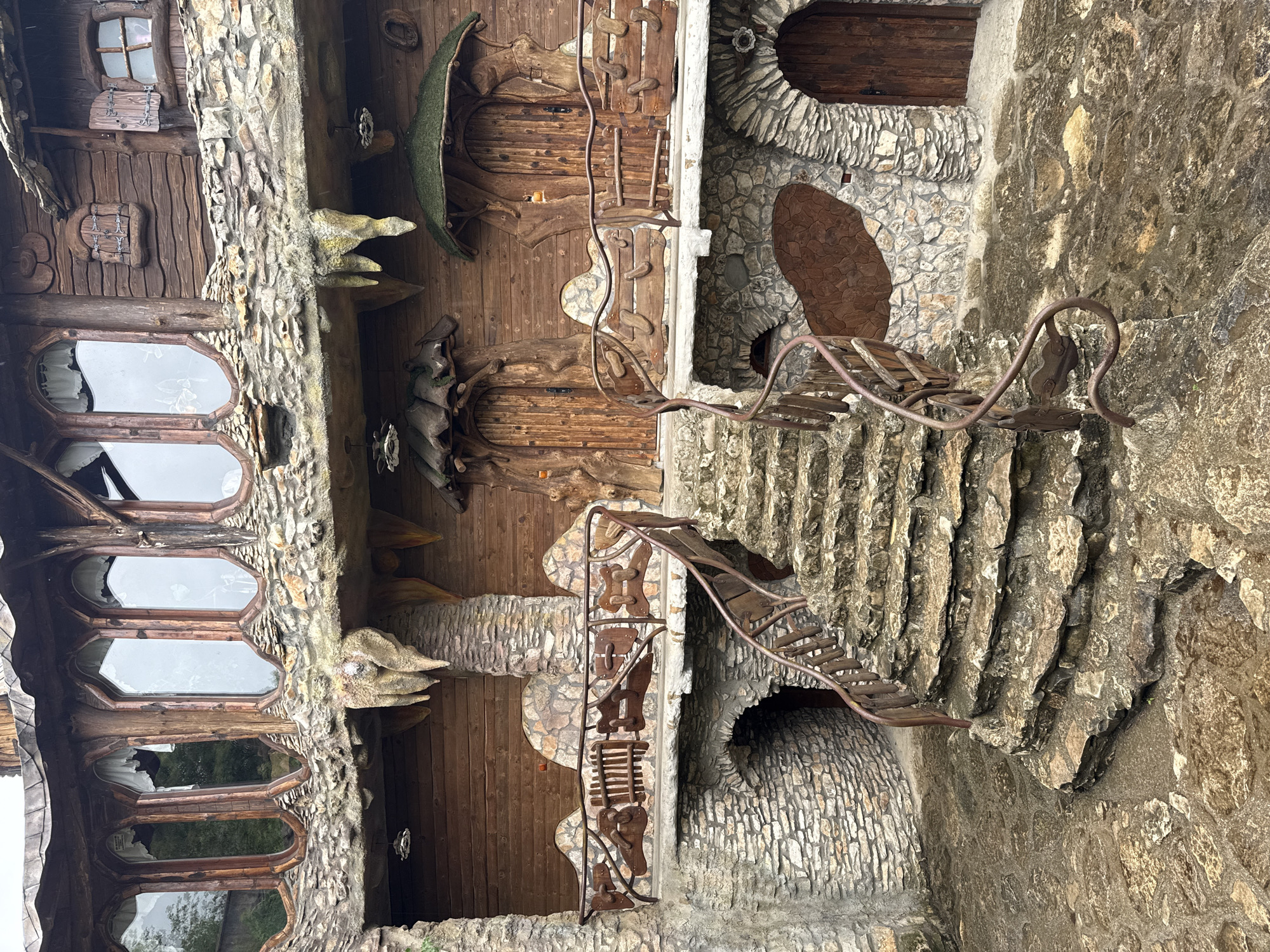 The image shows an exterior view of a structure, specifically the private house of sculptor Ali Mollov in Lukovit, Bulgaria. The architecture combines natural elements such as stone and wood, creating an organic, whimsical aesthetic. Notable features include irregularly shaped windows with wooden frames, intricately carved wooden doors and details, and stone masonry with textured surfaces. A metal staircase and decorative metalwork accent the exterior. The scene is captured in daylight, with a slight upward tilt, highlighting the architectural details of the facade.