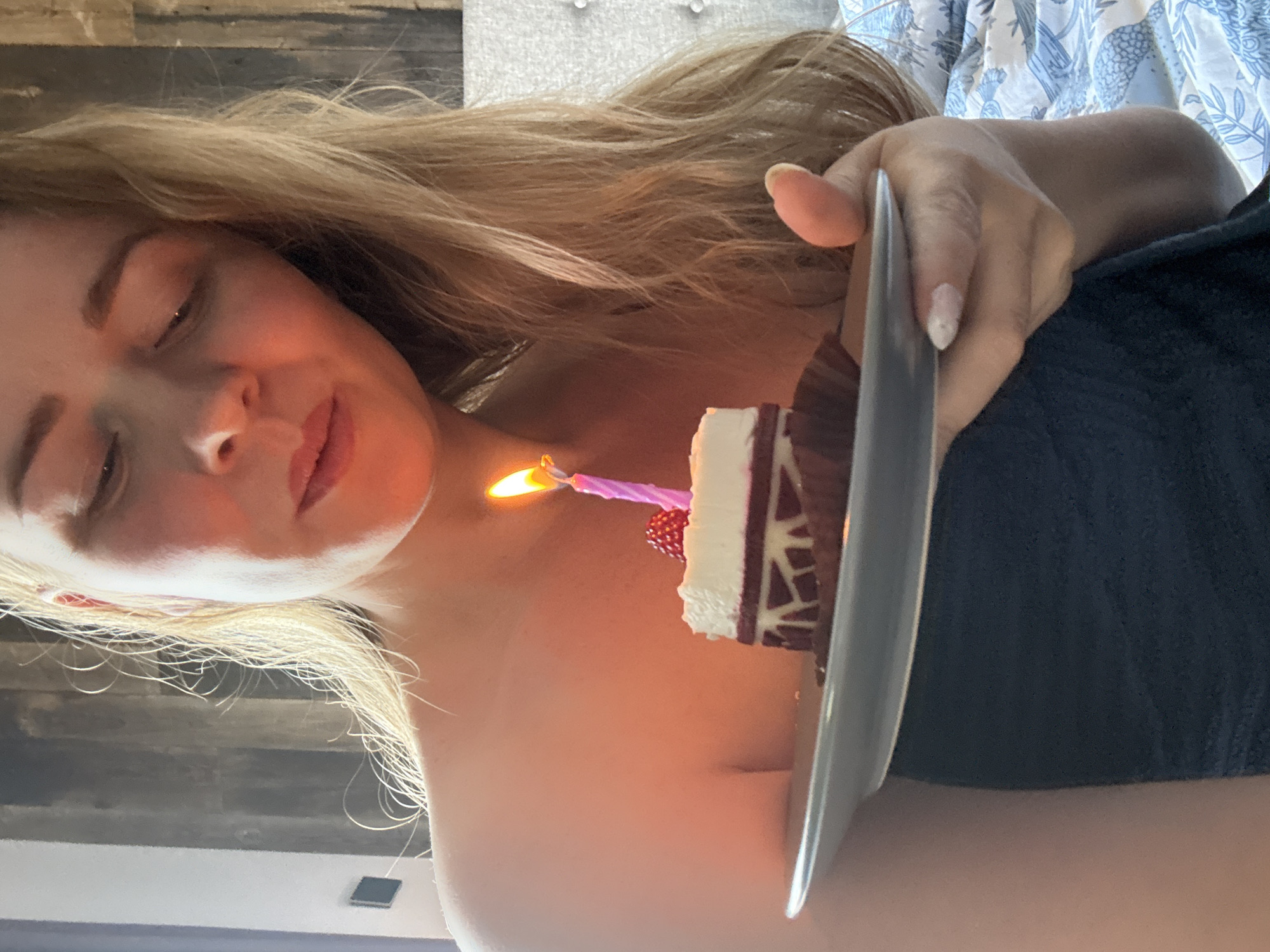 A woman with blonde hair is shown in a close-up shot, holding a small piece of cake on a plate. A lit candle is on the cake. She has her eyes closed and is slightly smiling. The woman is wearing a dark blue covering. The background includes a headboard and a bed with a blue and white patterned sheet. The location is not specified and there is no mention of Saint Petersburg, Russia.
