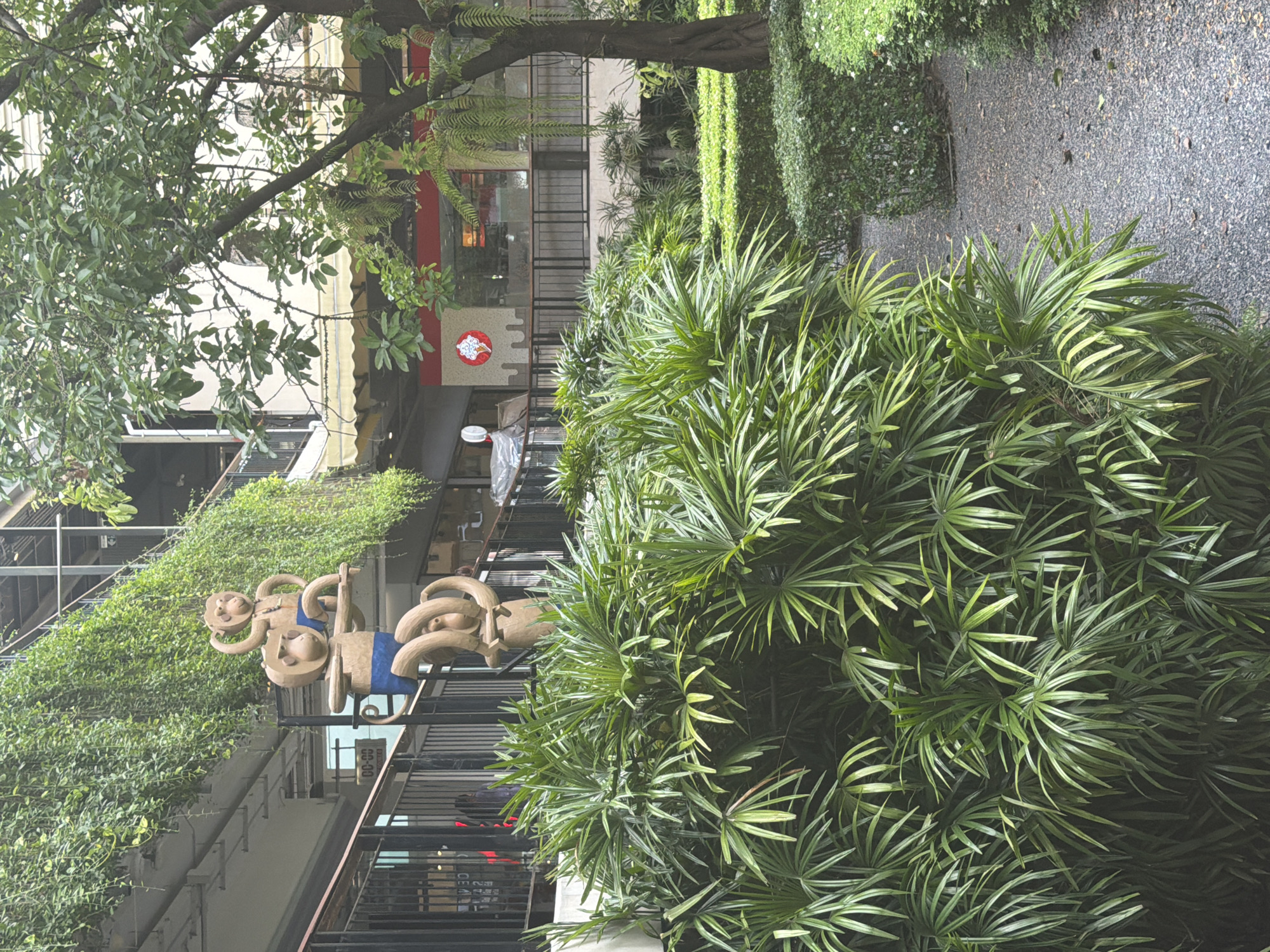 The image captures a scene in the city of บ้านบางรักษ์, Thailand. The foreground features dense green foliage of various plant species. A sculpture composed of light-brown figures with blue accents stands to the left, partially obscured by the foliage. In the background, buildings with balconies and open-air corridors are visible, displaying a mix of modern architecture and green walls with vegetation. A red sign with a white logo is visible above a storefront. A stone-paved walkway extends into the distance alongside the greenery. No people are distinctly visible in the scene.