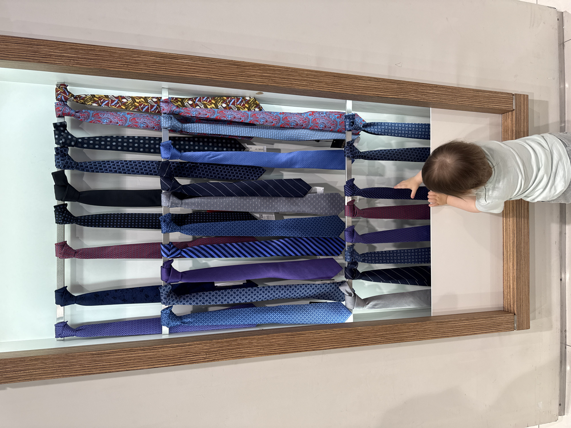 A top-down shot shows a child reaching into a drawer displaying numerous ties in a store. The ties are neatly arranged, hanging from what looks like metal rods. The drawer is framed with a wood-grained material, while the surrounding wall is a light color. The child is wearing light-colored clothing. The location is Khimki, Russia.