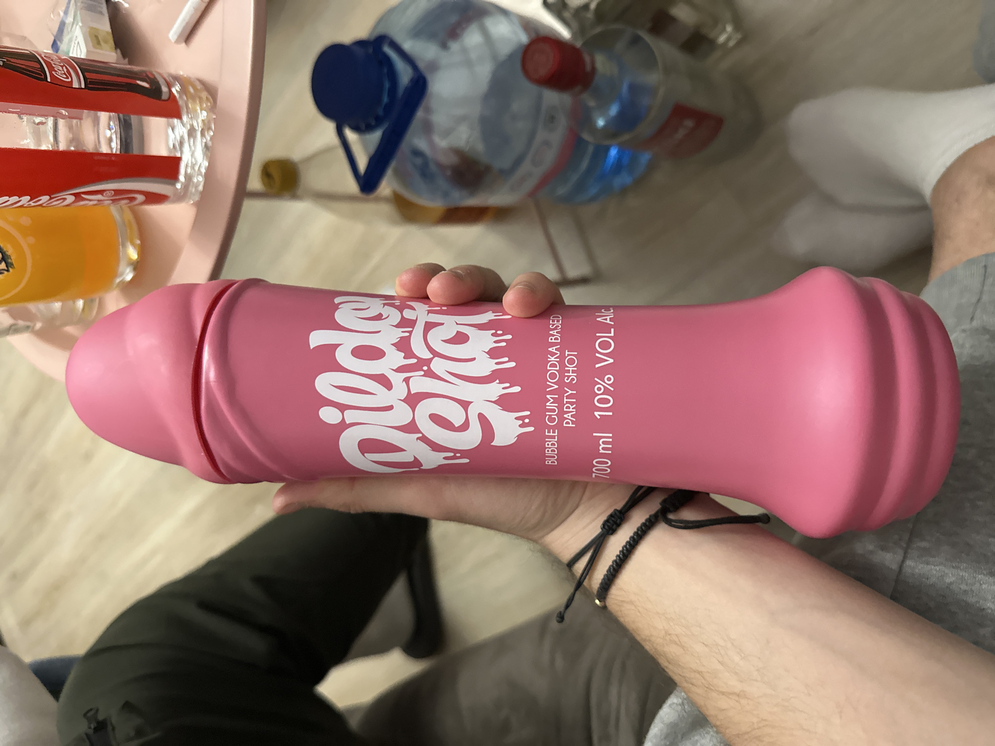 The image shows a person holding a pink, phallic-shaped bottle of "Dildo Shot" bubble gum vodka. The bottle is labeled with the brand name "Dildo Shot," "Bubble Gum Vodka Based Party Shot," and "700 ml 10% VOL Alc." The person is wearing grey sweatpants and a black bracelet. In the background, there are bottles of water, coca-cola and orange juice on a pink table. A floor is also visible. The image was not taken in Graf Ignatievo, Bulgaria.