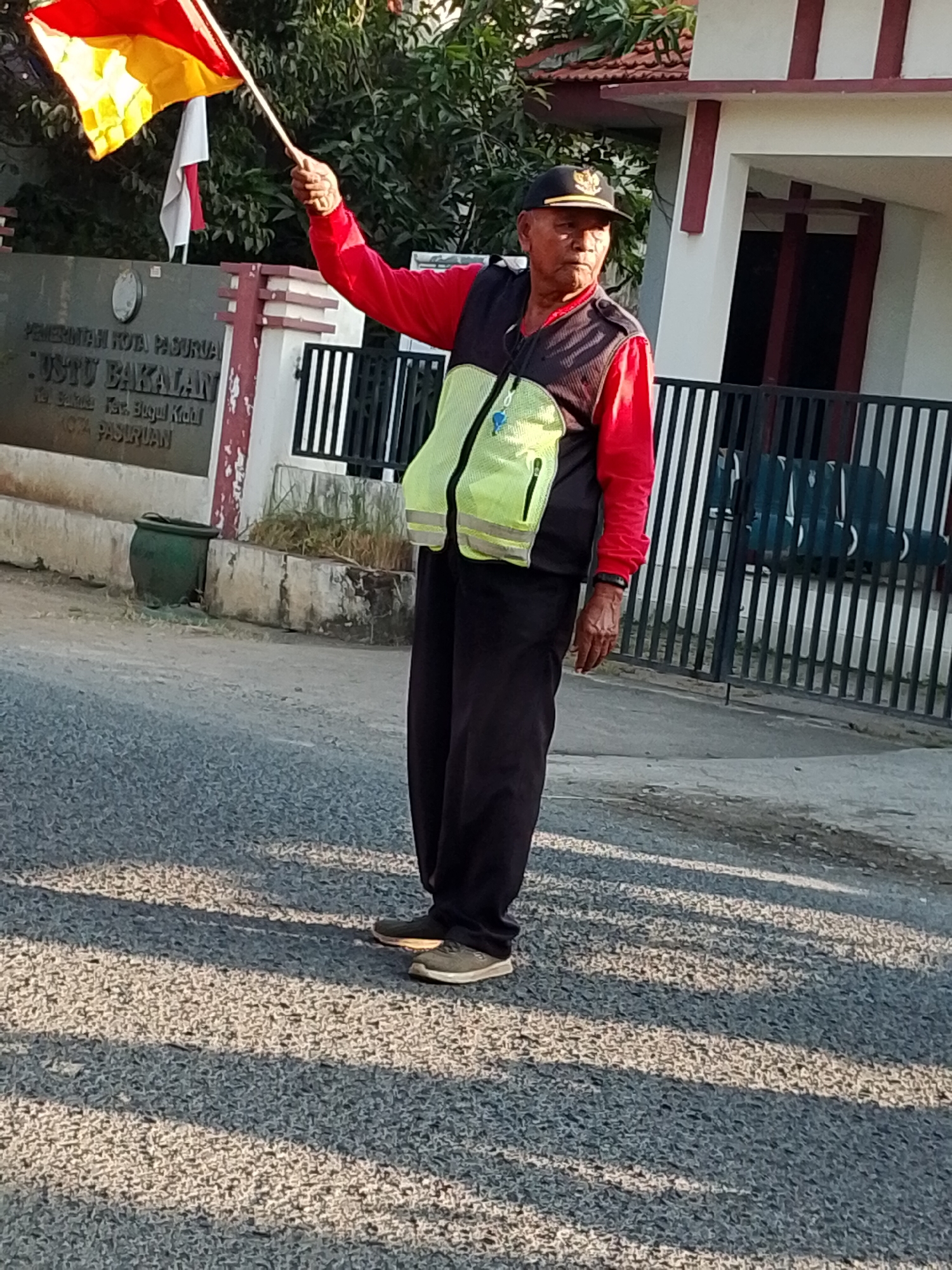 An elderly Indonesian man wearing a red long-sleeved shirt, a dark vest, a bright yellow reflective vest, and a black baseball cap is standing on a paved road, holding a red and yellow flag on a pole. He appears to be directing traffic or signaling. Behind him is a building with a sign that reads "PEMERINTAH KOTA PASURUAN" and "JALUR BAKULAN", indicating the location is in the city of Pasuruan, Indonesia. A black metal fence and some greenery are also visible in the background. The sun is casting long shadows on the road, suggesting it is either early morning or late afternoon.