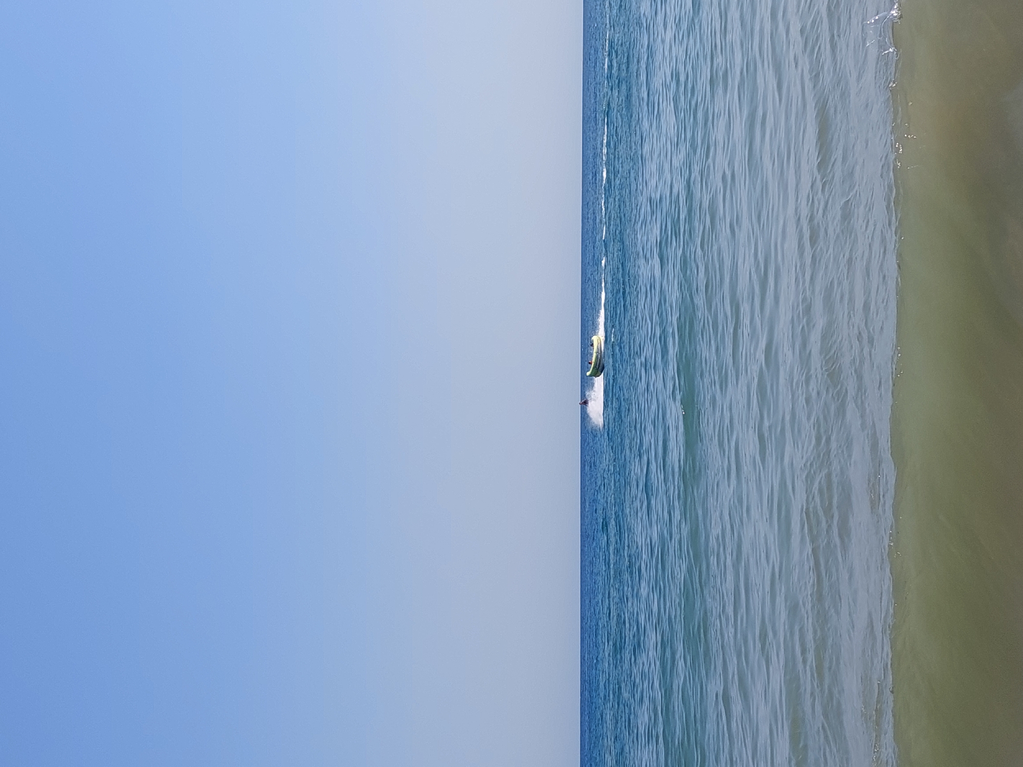 A horizontal, eye-level, full shot captures a bright, clear day at the Black Sea coast in Sveti Vlas, Bulgaria. The image is divided into three horizontal sections: a vast expanse of light blue sky, a band of deep blue sea with gentle waves, and a murky, light green shallow area in the foreground. In the middle ground, a yellow inflatable boat, possibly a "banana boat," is being pulled by an unseen boat, creating a white wake. A person is visible in the inflatable boat. The overall atmosphere is serene and summery.