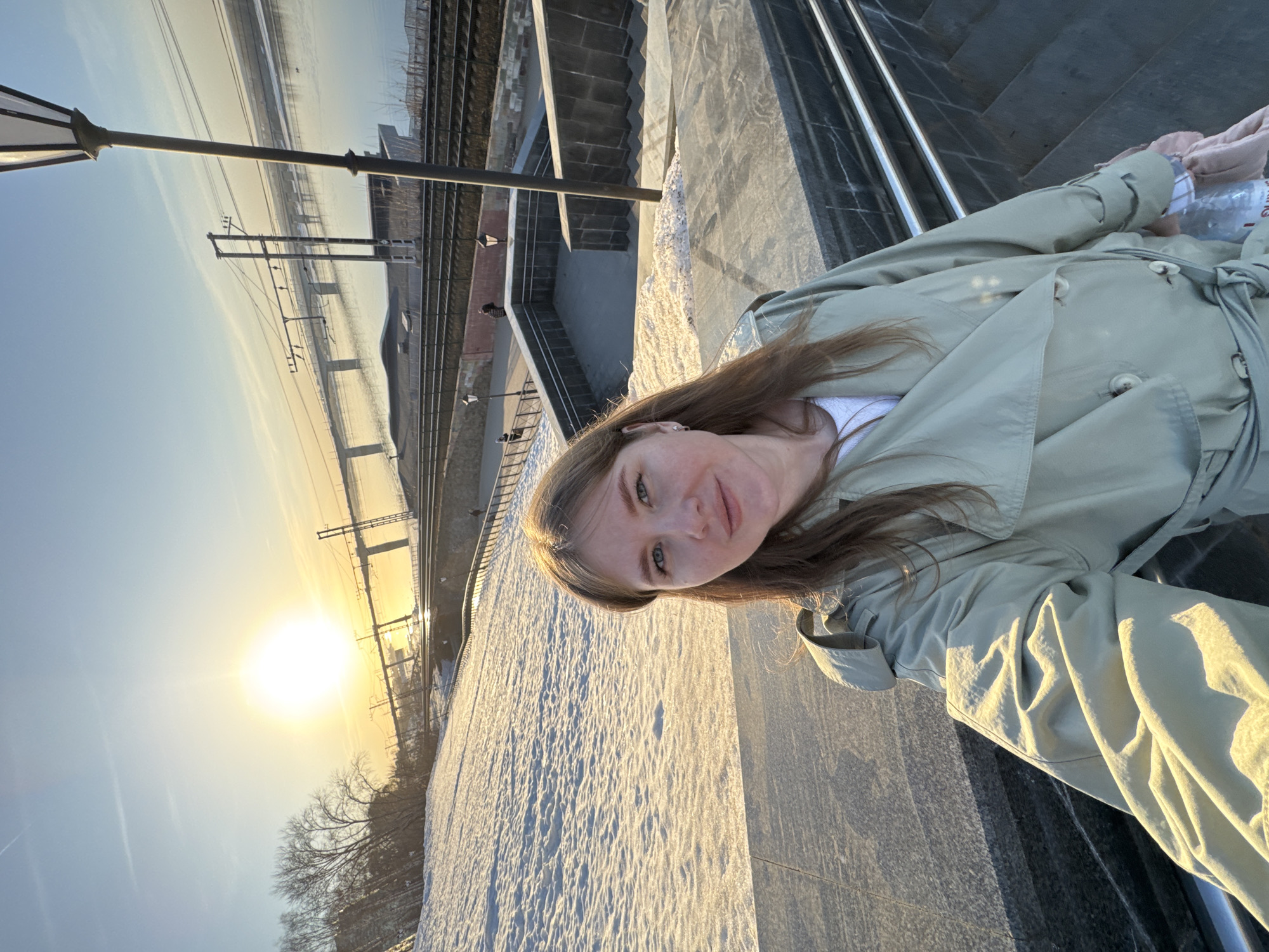 A woman with fair skin, light brown hair, and blue eyes is standing on a snow-covered surface, likely a walkway or platform. She is wearing a light green trench coat and a white shirt underneath. The background shows a landscape with railroad tracks, a bridge, and bare trees silhouetted against the bright sun. The light suggests it is either sunrise or sunset. The location appears to be a viewpoint overlooking the Kama River in Perm, Russia, based on the presence of the railway infrastructure and the characteristic river landscape. In her hand, she holds a plastic bottle of water.