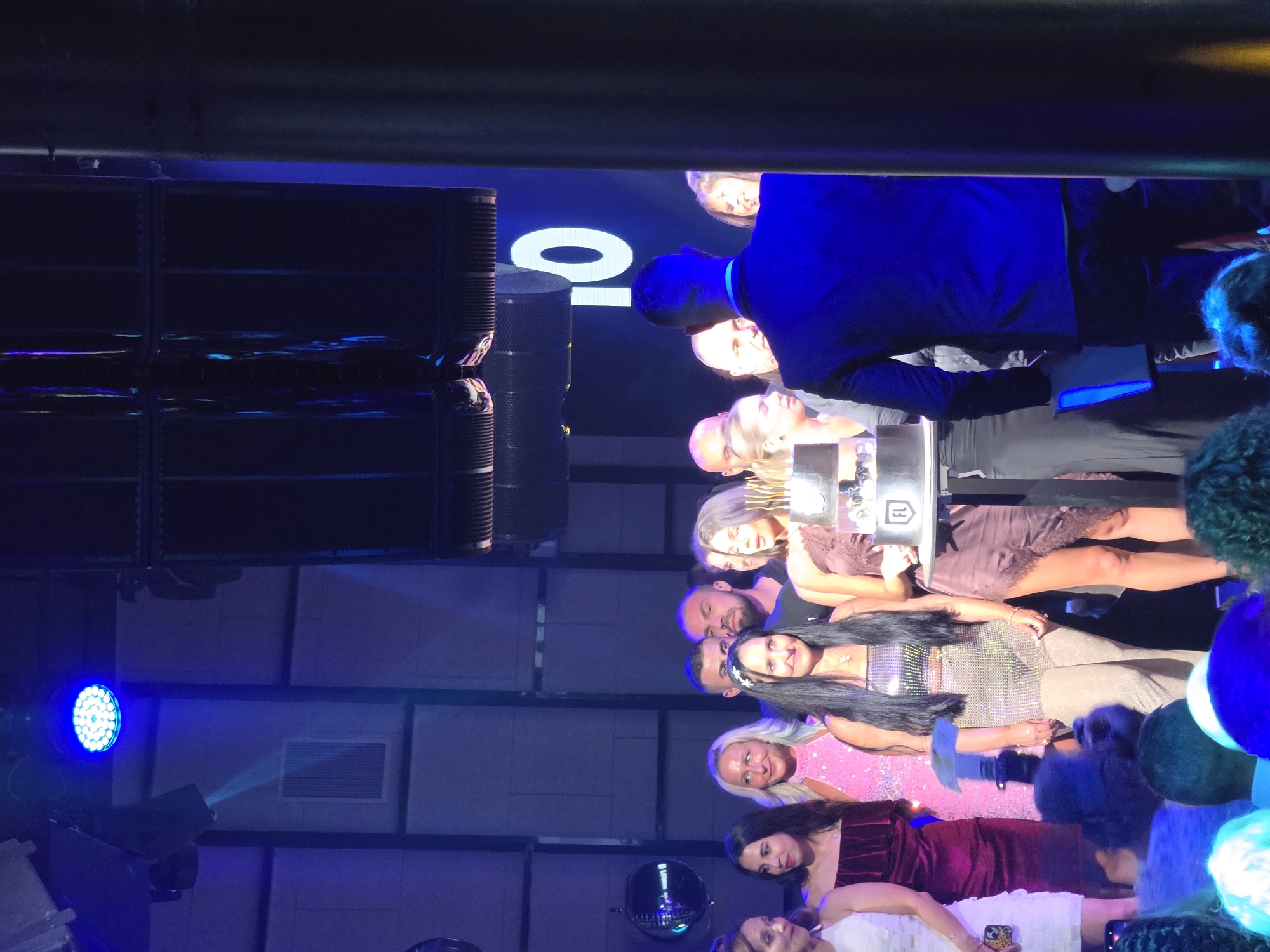 An outdoor, nighttime birthday celebration is taking place, featuring a group of people gathered around a tiered cake. The background displays a large screen with the number "10" illuminated in white. The event appears to be on a stage or platform, with blue lighting casting a dramatic effect on the scene and the surrounding structures. Some attendees are dressed in festive attire, with one woman wearing a sparkly outfit and another in a maroon dress. The mood seems celebratory, with a cake prominently featured as the centerpiece of the event. The specific location is the Varna City Center in Varna, Bulgaria.
