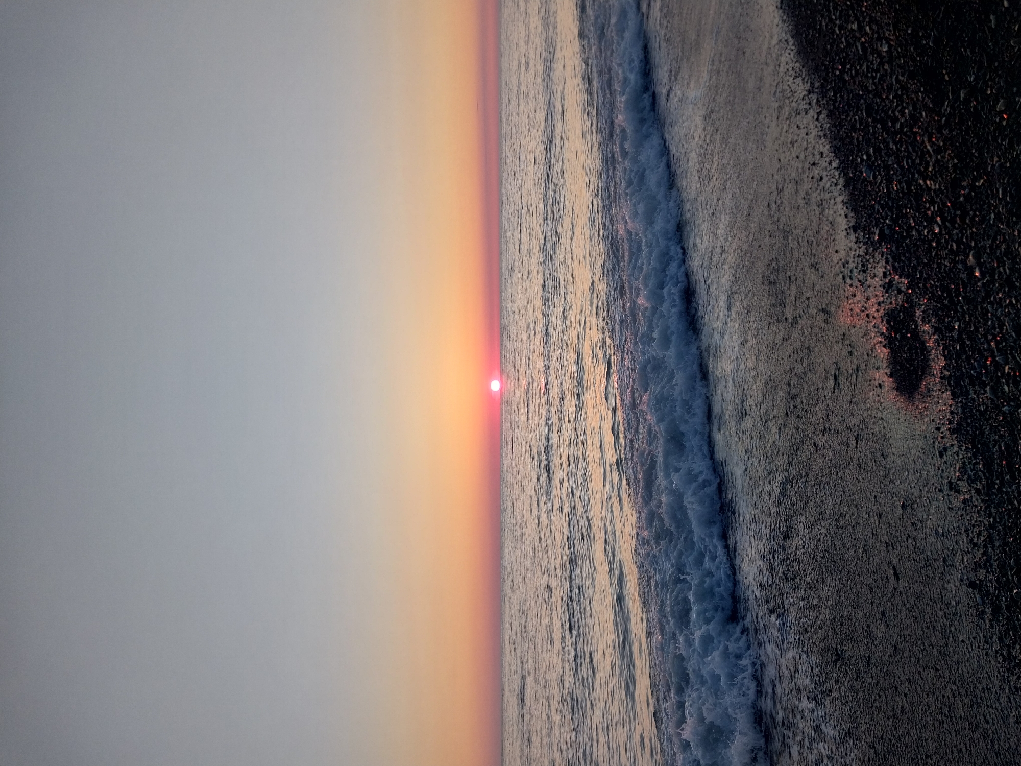 This media file captures a serene coastal landscape at either sunrise or sunset, set in Sochi, Russia. The image is oriented horizontally, showcasing a clear horizon line that separates the sky from the sea.
The top portion of the frame is dominated by the sky, which exhibits a beautiful gradient. It transitions from a soft, light grey-blue at the very top to a warm, ethereal blend of orange and subtle pink hues as it approaches the horizon. A small, brilliant red sun is perfectly centered on the horizon line, marking the low position of the sun.
Below the horizon, the expansive sea fills the middle ground. Its surface reflects the warm colors of the sky, creating shimmering patterns on the water. A prominent wave is captured in motion, actively breaking and receding towards the foreground. This wave displays frothy white foam along its crest and turbulent, blue-grey water as it washes onto the shore.
The bottom section of the photograph reveals a dark, pebble beach, wet from the receding tide. The damp pebbles subtly reflect faint reddish light from the sun, adding depth to the foreground. No individuals or man-made structures are visible, maintaining the pristine and natural essence of the scene. The overall impression is one of tranquil beauty and the quiet spectacle of a coastal dawn or dusk. There is no visible text in the image.