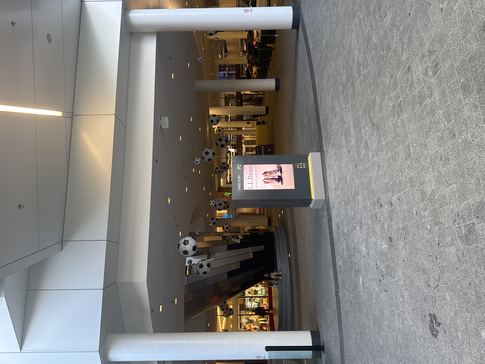 An outdoor pedestrian area in front of a building appears to be a mall entrance or plaza. The ground is paved with grey square tiles. Several large, black and white soccer ball-shaped decorations are suspended from the ceiling by thin wires. In the midground, a woman is sitting on a curved, tiered staircase with a dog next to her. Behind them, store windows display mannequins. To the right of the staircase is a digital directory sign with "LL Bean" prominently displayed. Beyond the directory sign, several cars are parked. The architecture features large white columns and a modern, angular facade. The scene suggests a retail or entertainment district, likely in an urban setting. The location is Brentwood Town Centre, Burnaby, Canada.