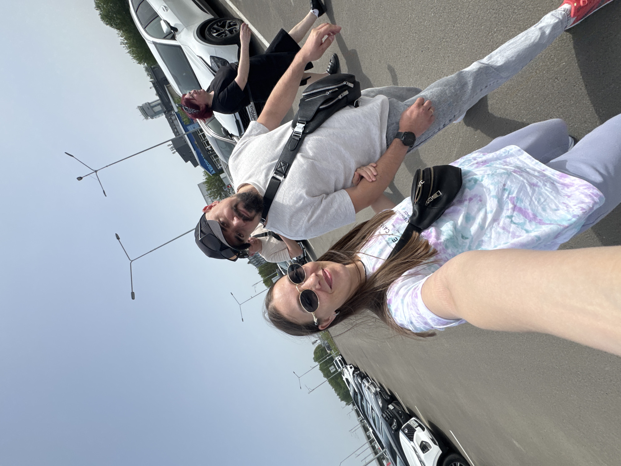 The photograph shows a group of people posing for a selfie outdoors, presumably in Bolshiye Kabany, Russia, though specific landmarks are not discernible. A woman holds the camera; she wears sunglasses and a patterned t-shirt. A man in a baseball cap and light t-shirt stands behind her. Another person, with reddish hair, appears to be lying on the hood of a white car in the background. Several cars are parked along the edge of the frame, suggesting a parking lot or roadside location. Streetlights are visible in the background. The overall impression is a casual, informal moment captured in a sunny outdoor setting.